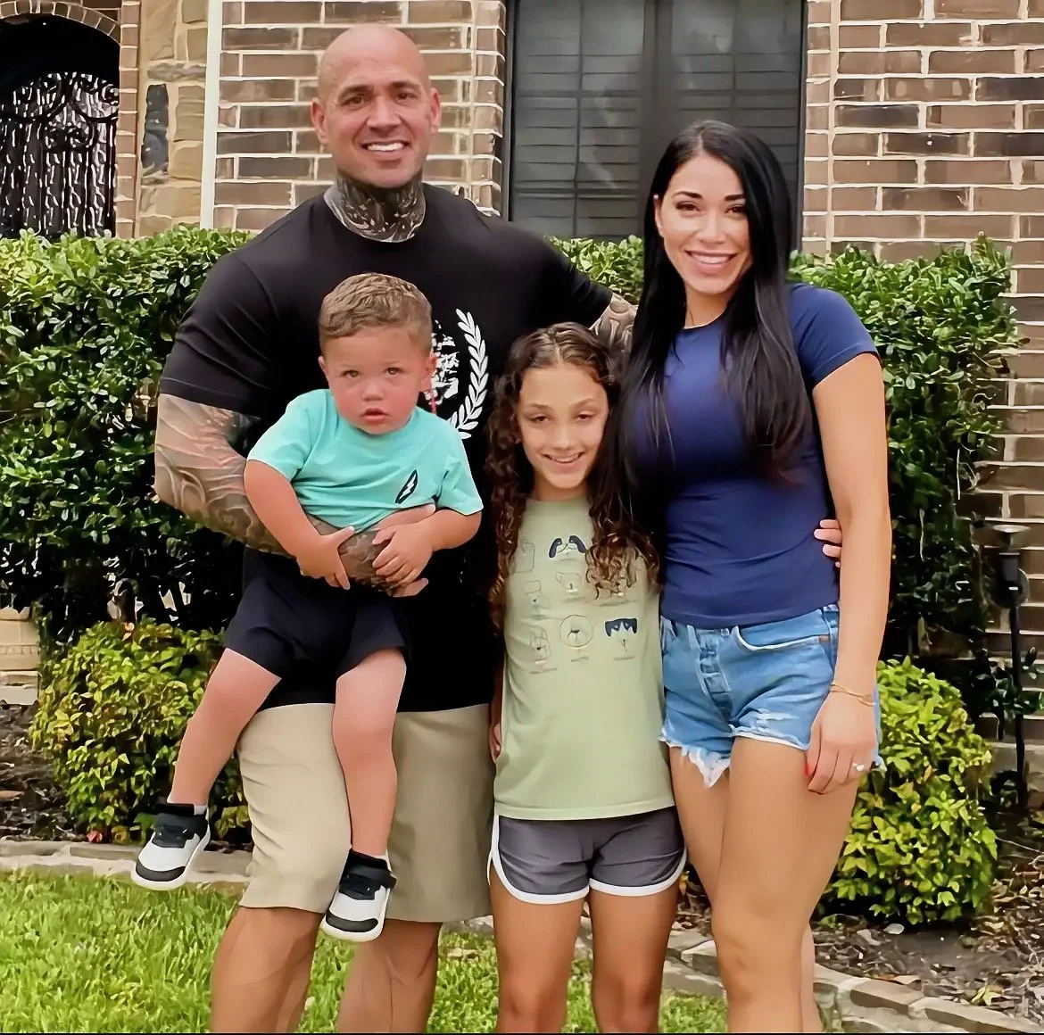 A smiling family of four standing outside in front of a brick house and greenery. The father is holding a young boy, and a woman with long dark hair stands next to a girl with curly hair.