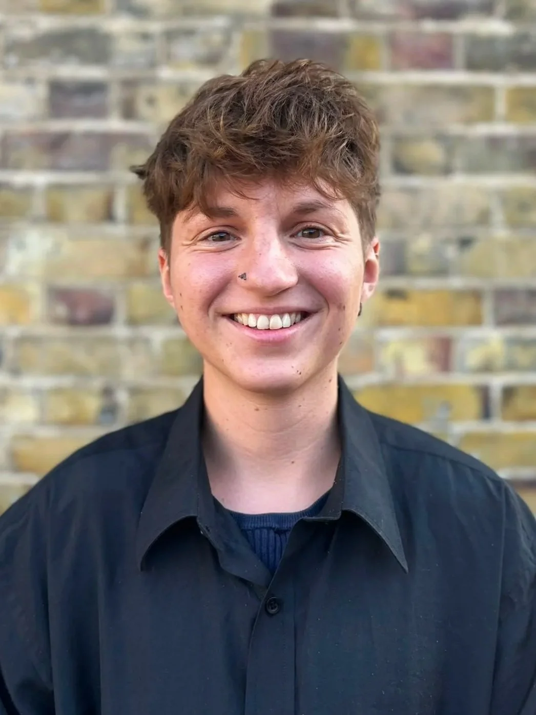 Young man with brown hair, smiling, wearing a black shirt, standing against a blurred brick wall background.