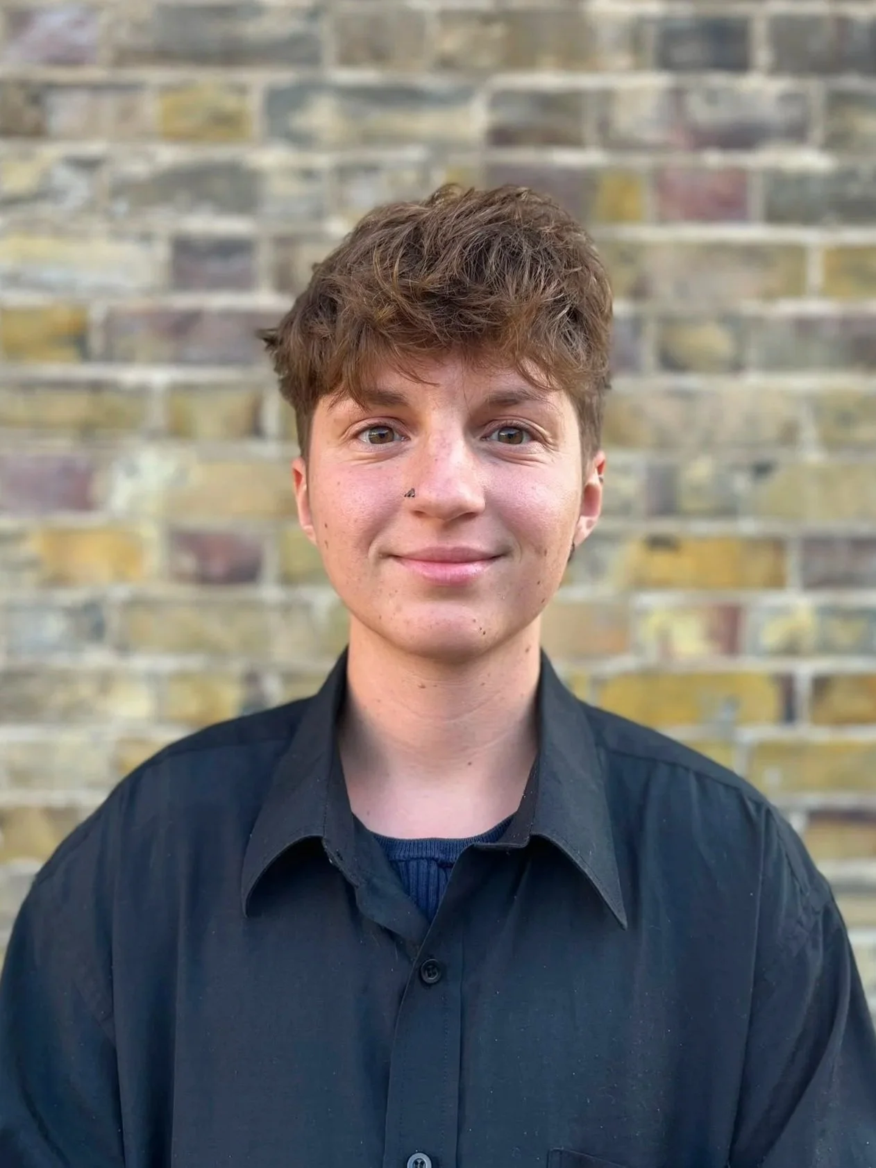 A young person with short, curly brown hair, wearing a black button-up shirt, standing in front of a brick wall.