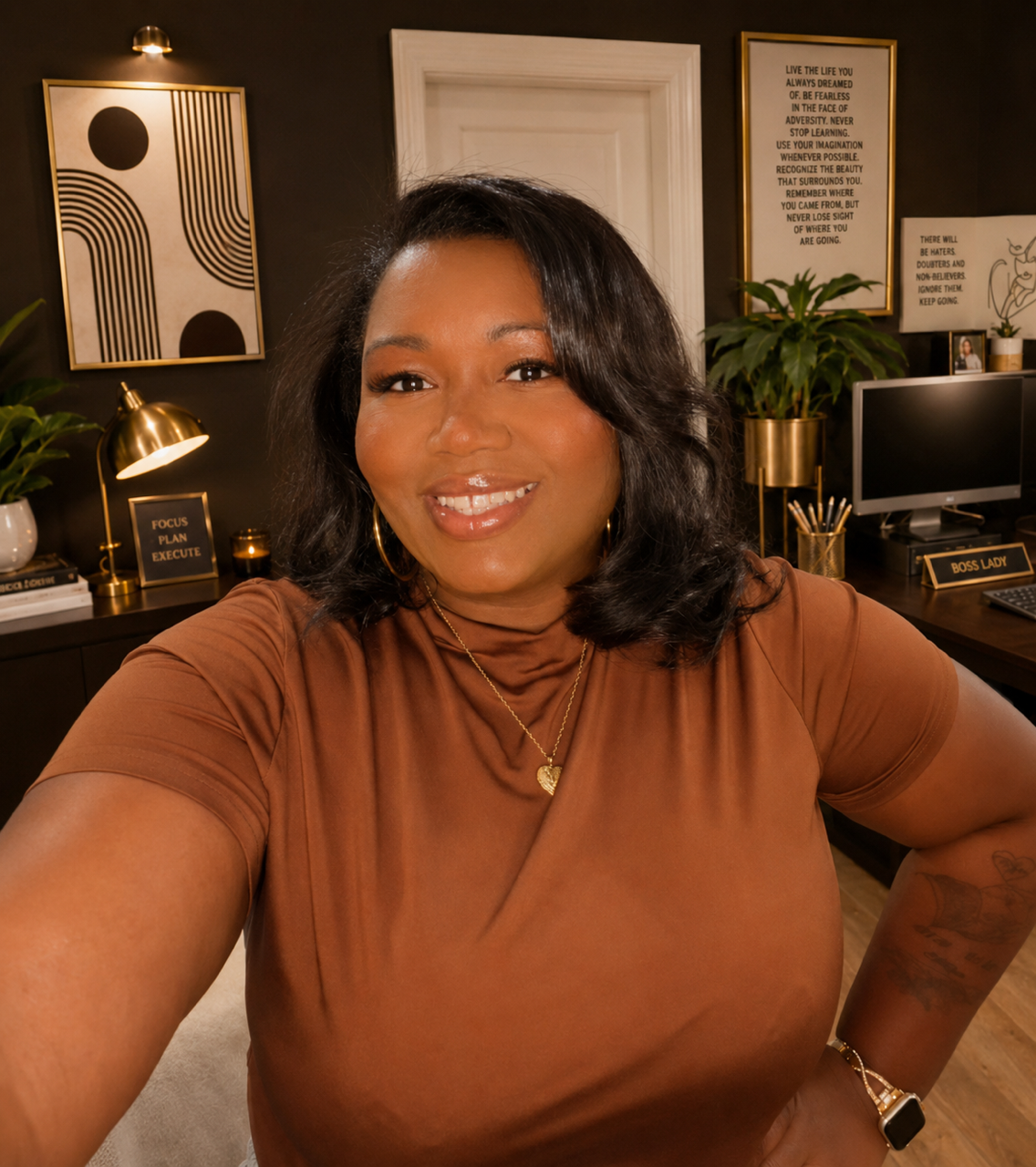 A woman with dark hair and wearing a brown top taking a selfie in an office. The office has motivational and inspirational wall art, a computer, plants, and desk accessories.
