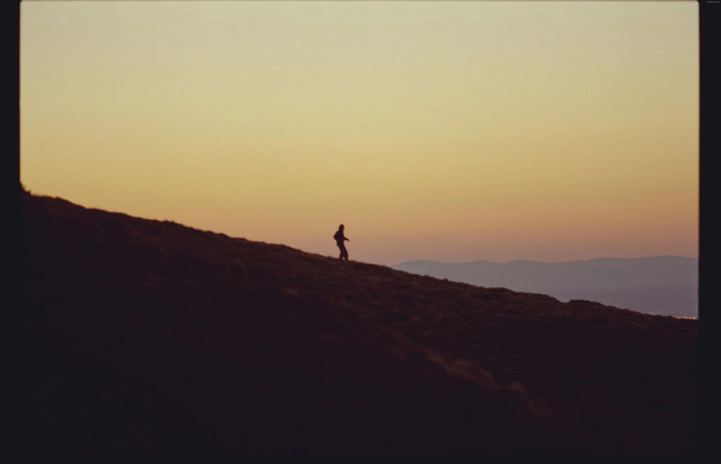 Silhouette of a person walking on a hillside during sunset or sunrise with mountains in the background.