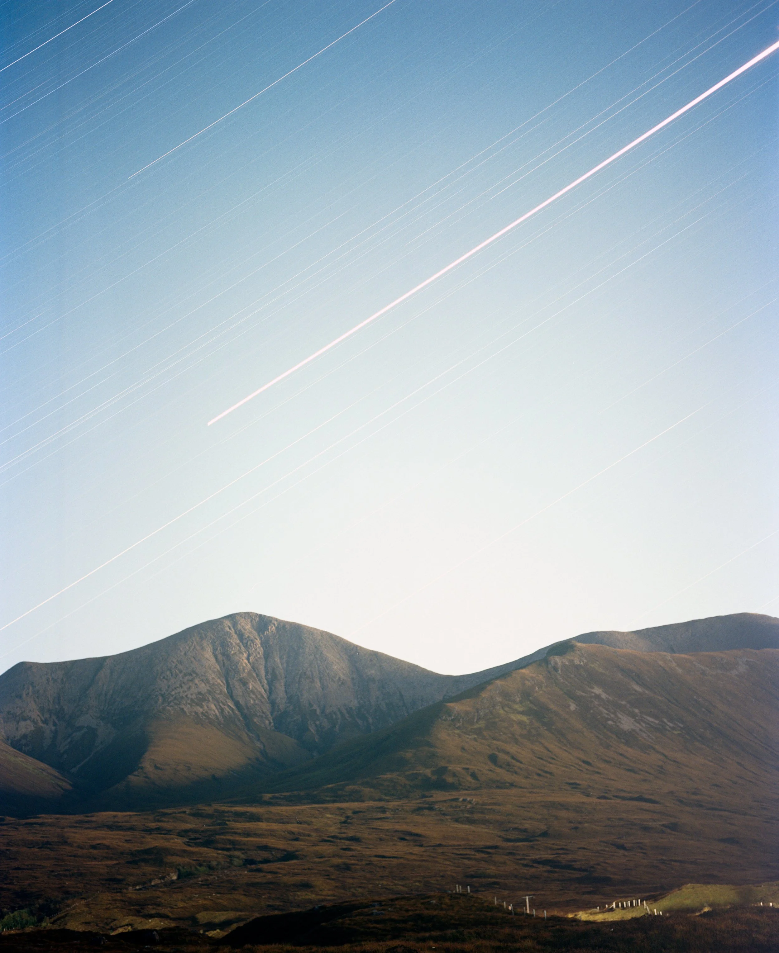 Night sky with star trails over a mountain landscape with rolling hills and a fence in the foreground.