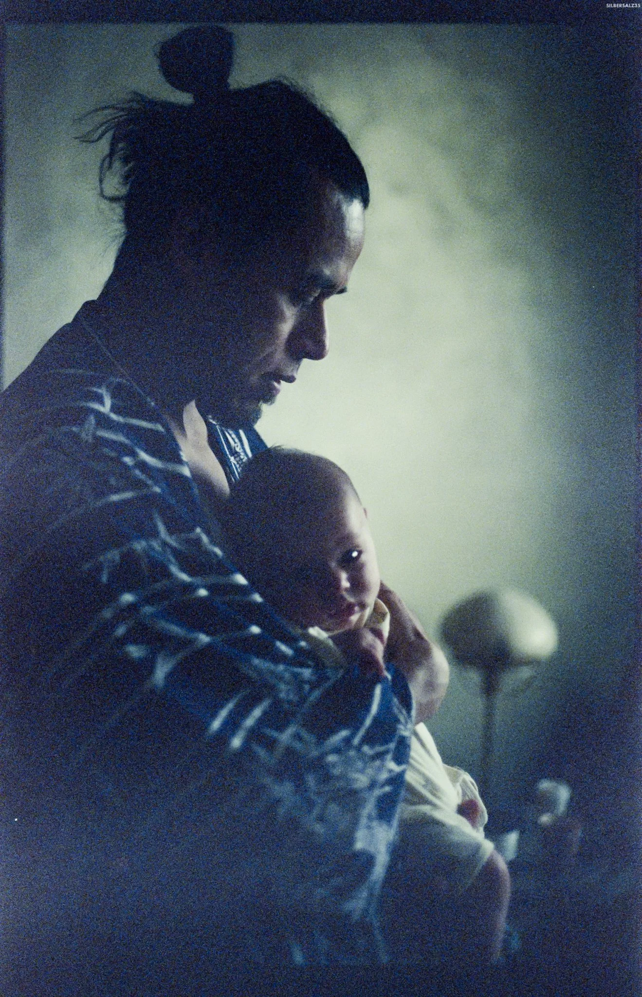 A man with braided hair holding a young child, both looking downward, with dim lighting and blurred background.