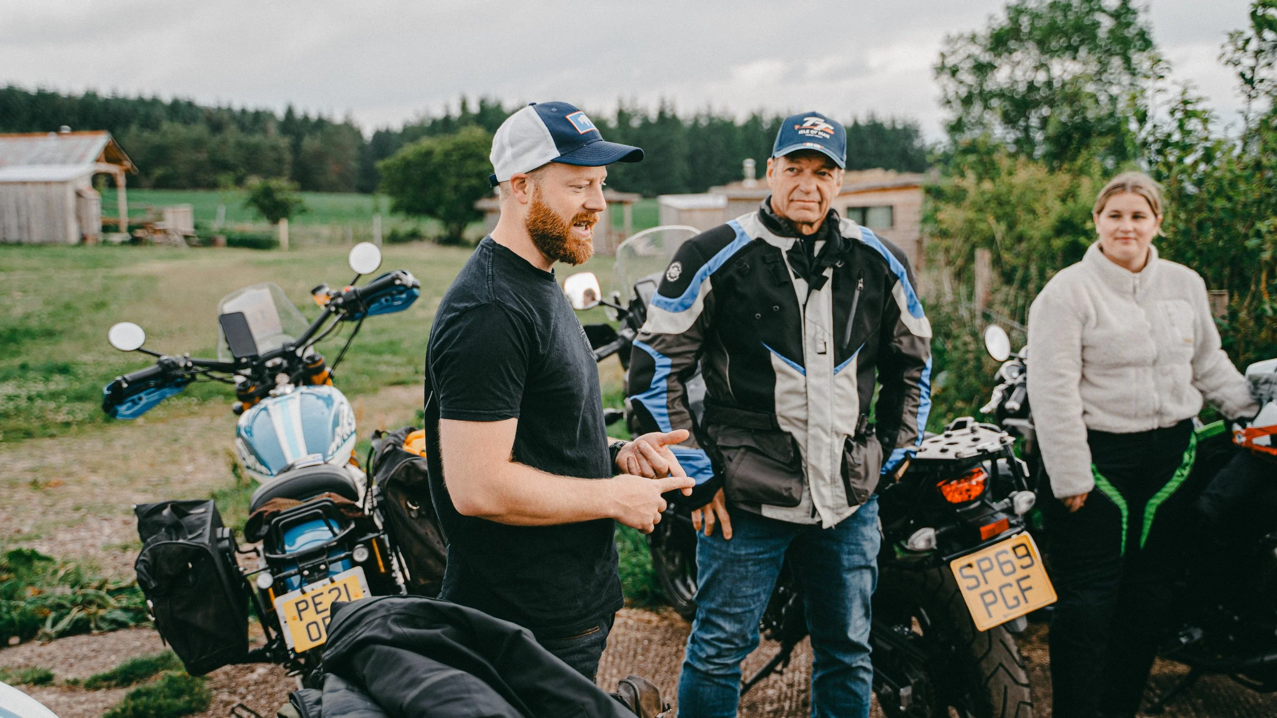 Three people standing next to motorcycles outdoors in a rural area, with green fields and trees in the background. The person on the left is talking, the middle person is wearing a motorcycle jacket and cap, and the third person is a woman in a light