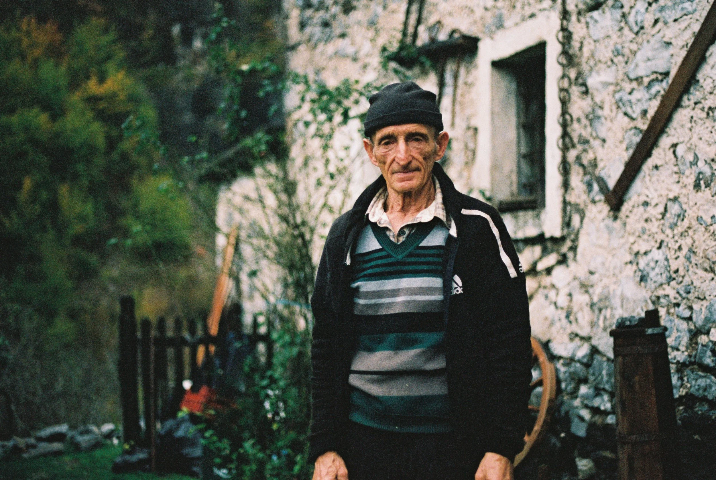 An elderly man wearing a black beanie, a striped shirt, and a black jacket walking outdoors near a stone building with small windows, surrounded by greenery.