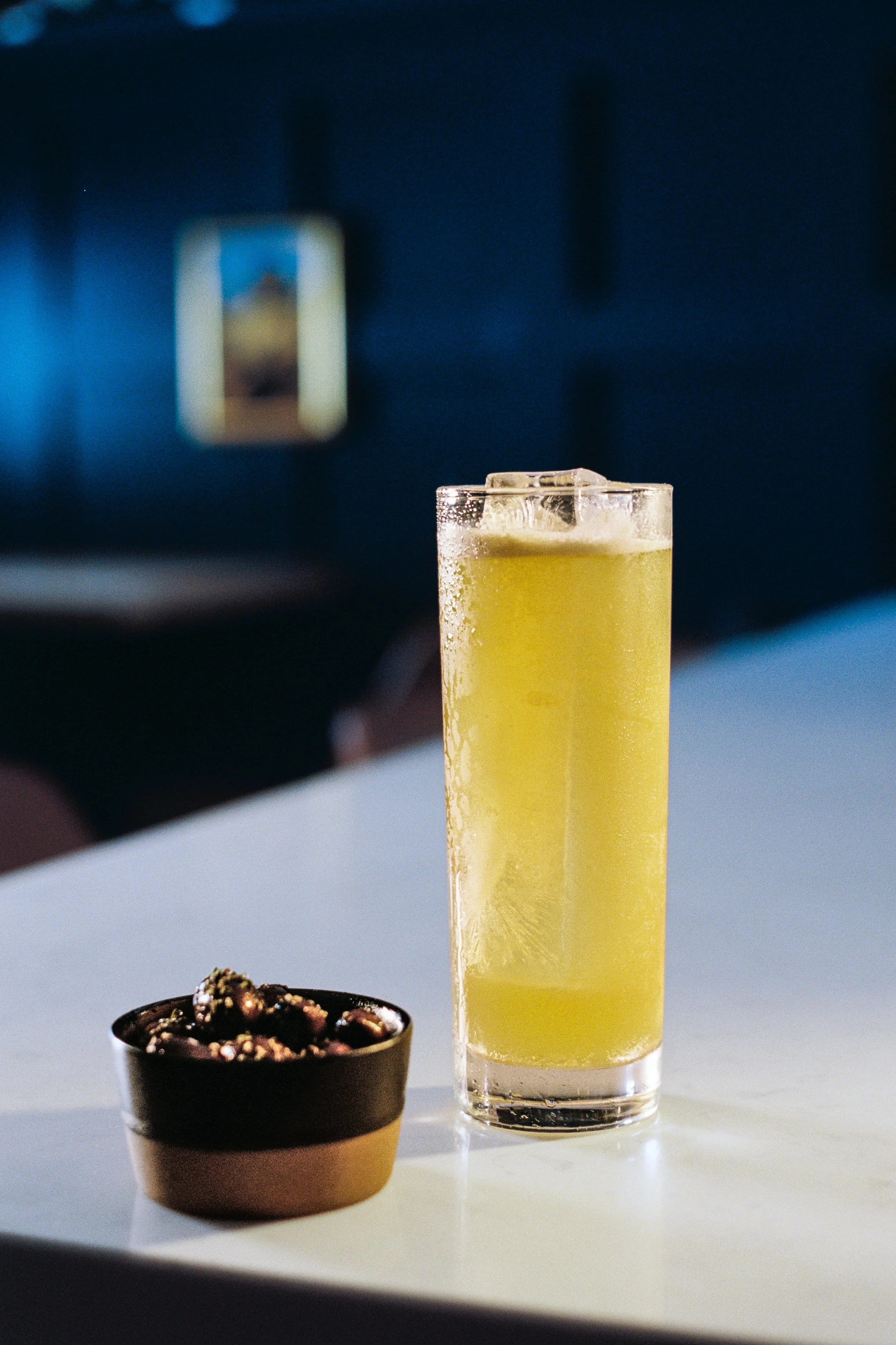 A tall glass filled with a yellow beverage and ice cubes, placed on a white surface, with a small bowl of chocolates or candies nearby and a blurred background featuring a rectangular object.