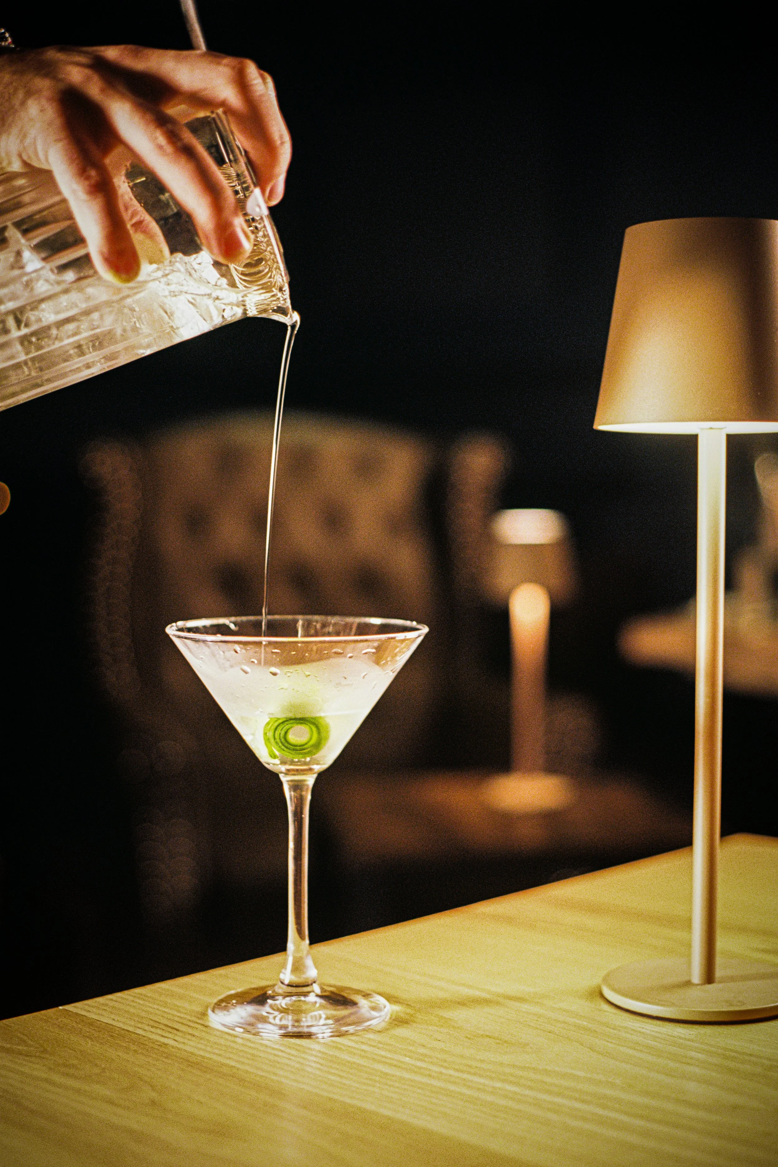A hand pouring clear liquid into a martini glass with a green olive inside, on a wooden table, with a lit gold-colored lamp nearby in a dimly lit setting.