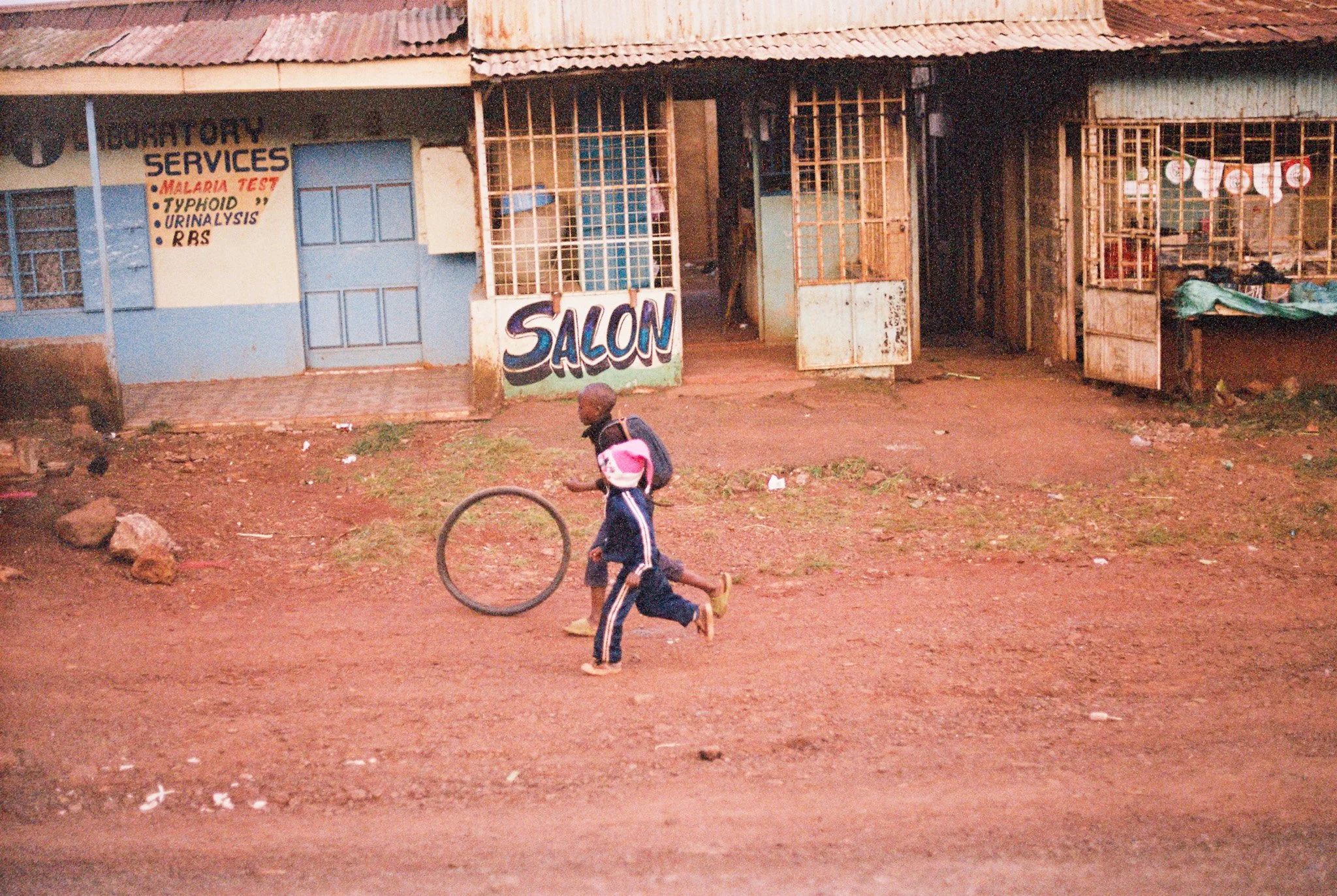A boy running on a dirt street in front of a building with signs advertising laundry and medical services and a salon.