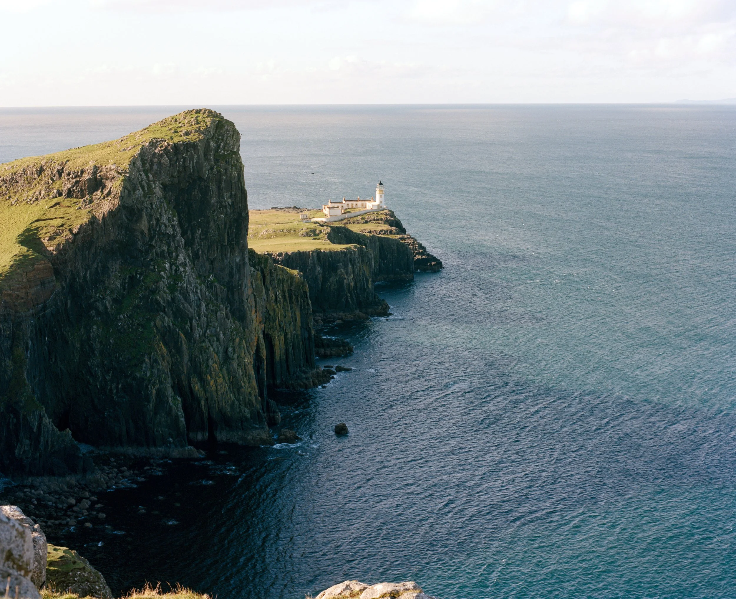 A lighthouse situated on a rocky green cliff overlooking the ocean with a cloudy sky overhead.