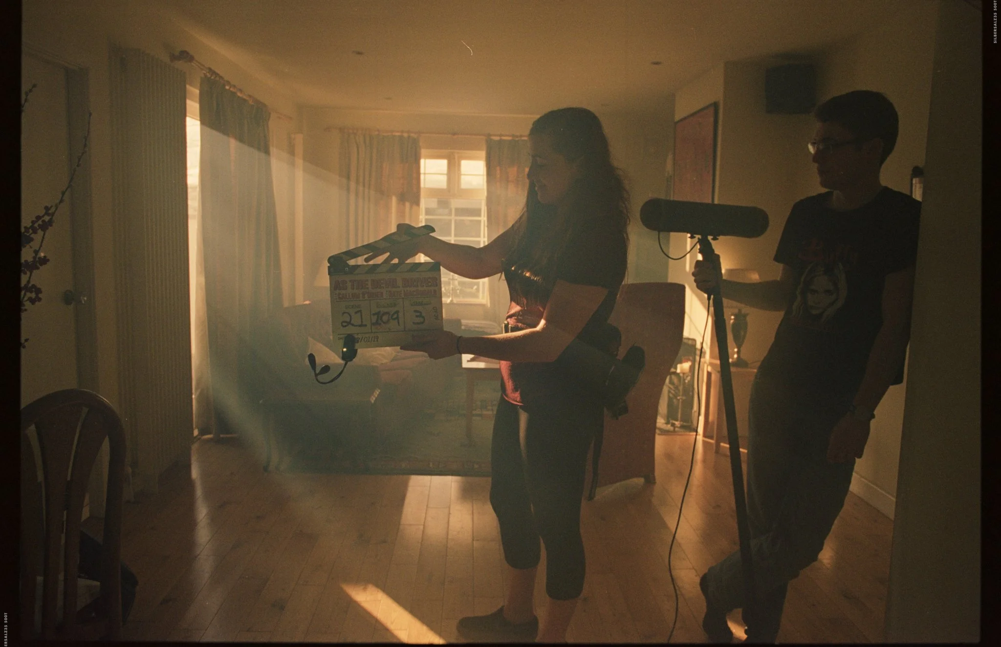 Two people on a film set handle a clapperboard, with sunlight streaming in through the windows of a living room, creating a backlit silhouette effect.