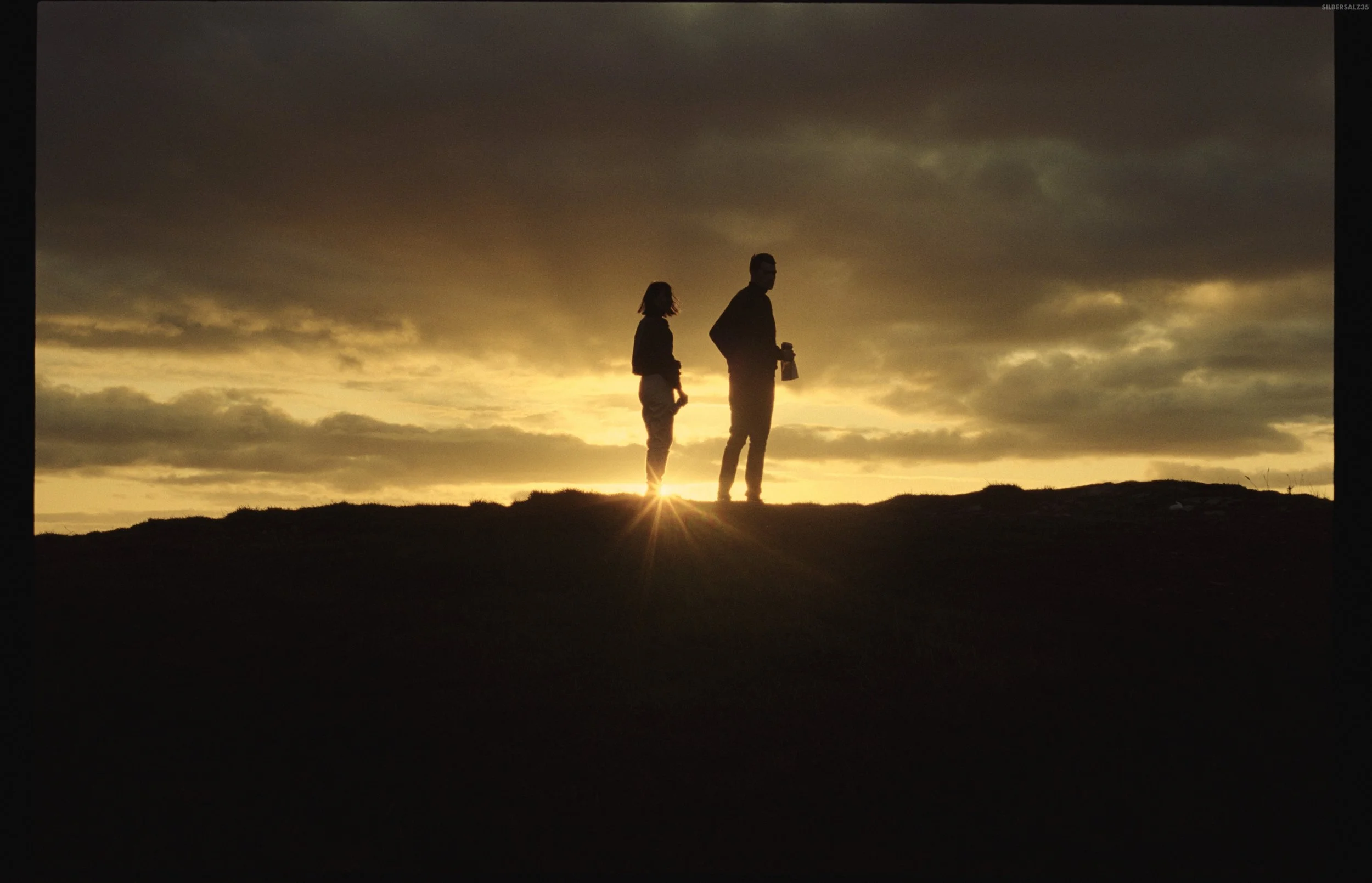 Silhouettes of a man and a woman standing on a hill at sunset, with the sun partially visible, casting a warm glow and creating long shadows.