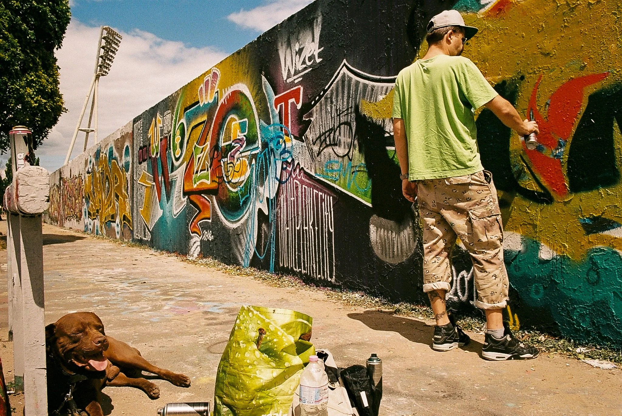 A man wearing a green t-shirt, camouflage shorts, black sneakers, and a grey cap standing in front of a colorful graffiti mural. A brown dog, lying on the ground with its tongue out, and various spray paint cans and bags are beside him. A sandy path 