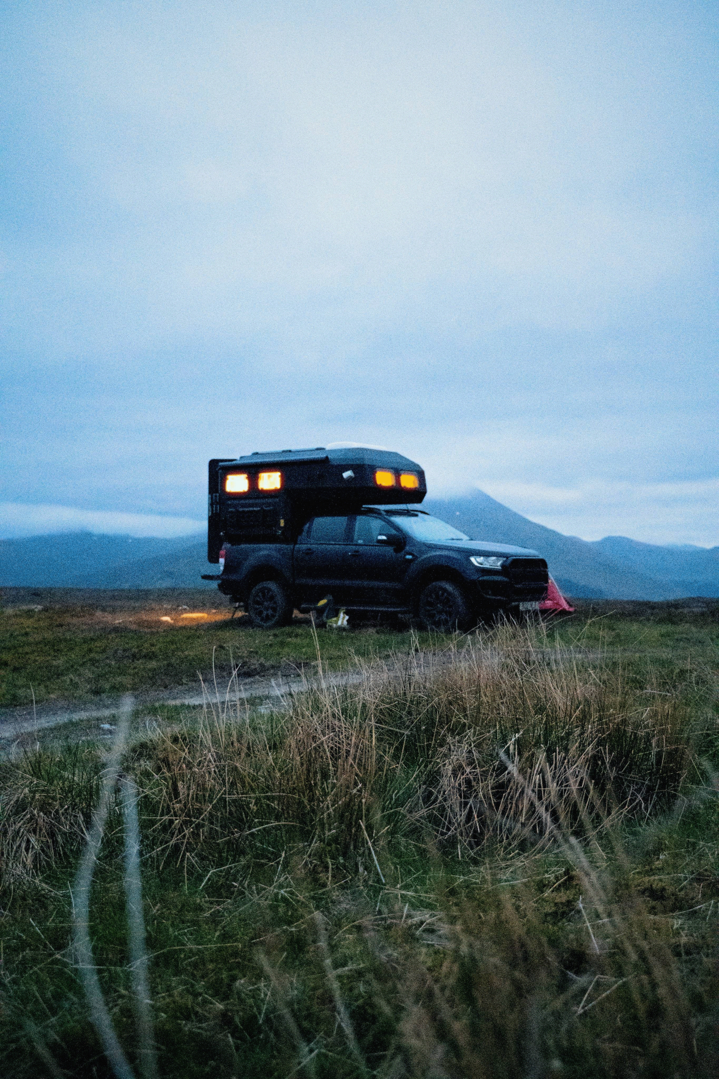 A black pickup truck with a rooftop camper parked on grassy terrain with mountains in the background during twilight.