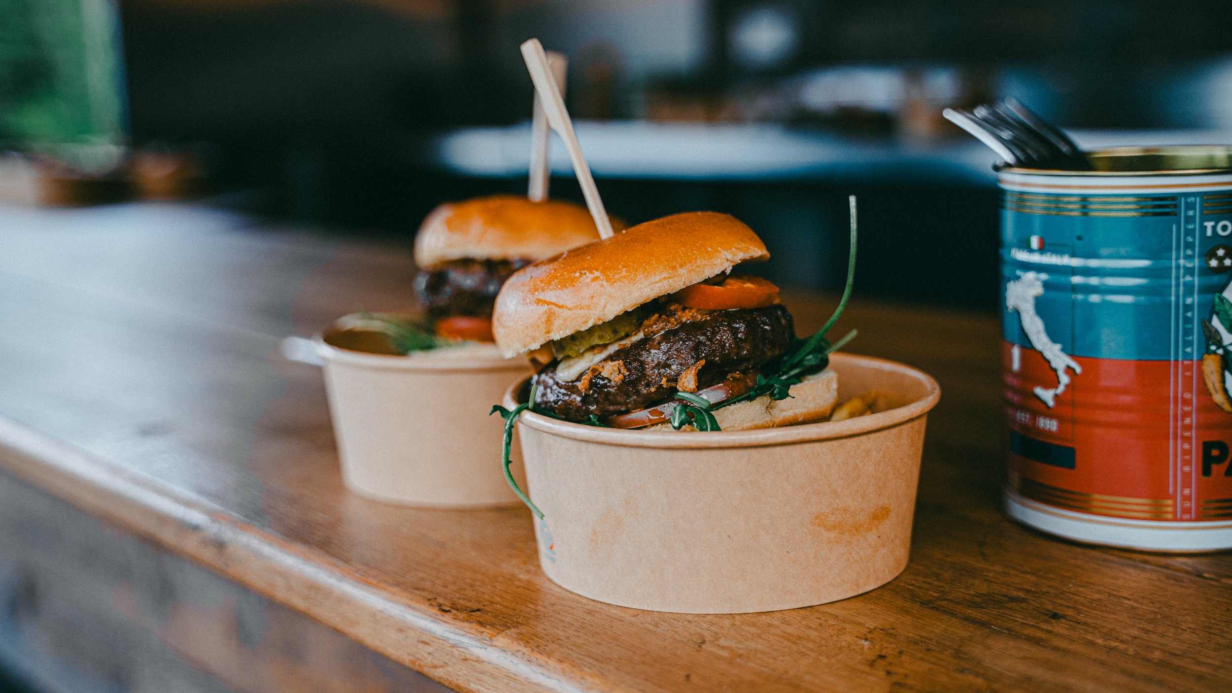 Two beef hamburgers with tomatoes, pickles, and greens in paper containers on a wooden surface, next to a canned drink with a straw.