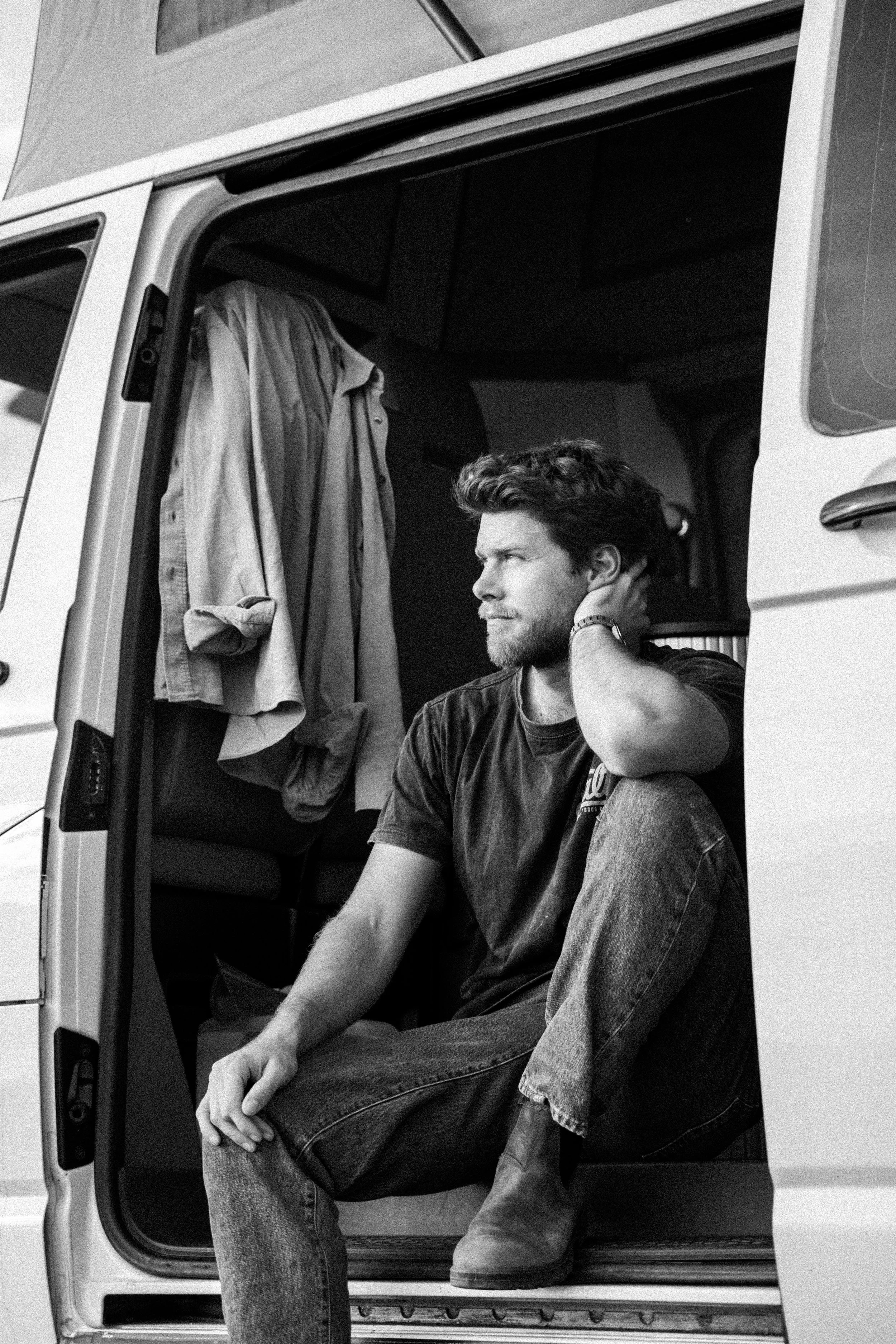 A black-and-white photo of a man sitting in the open doorway of a vehicle, with a shirt hanging inside the vehicle. The man is touching his ear or neck and looking to his left, wearing a dark t-shirt and jeans.