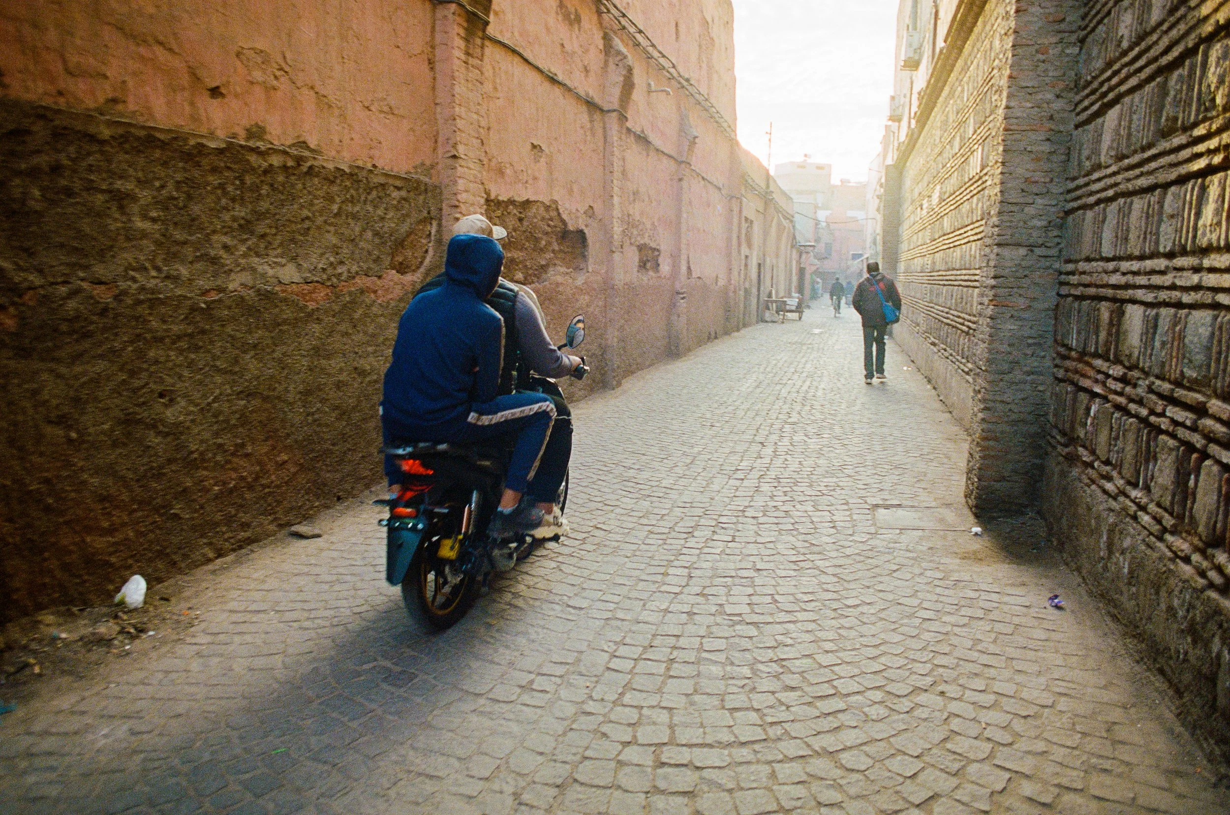 Two people riding a bicycle down a narrow cobblestone alleyway with brick and stucco walls on either side, and other pedestrians walking further ahead.