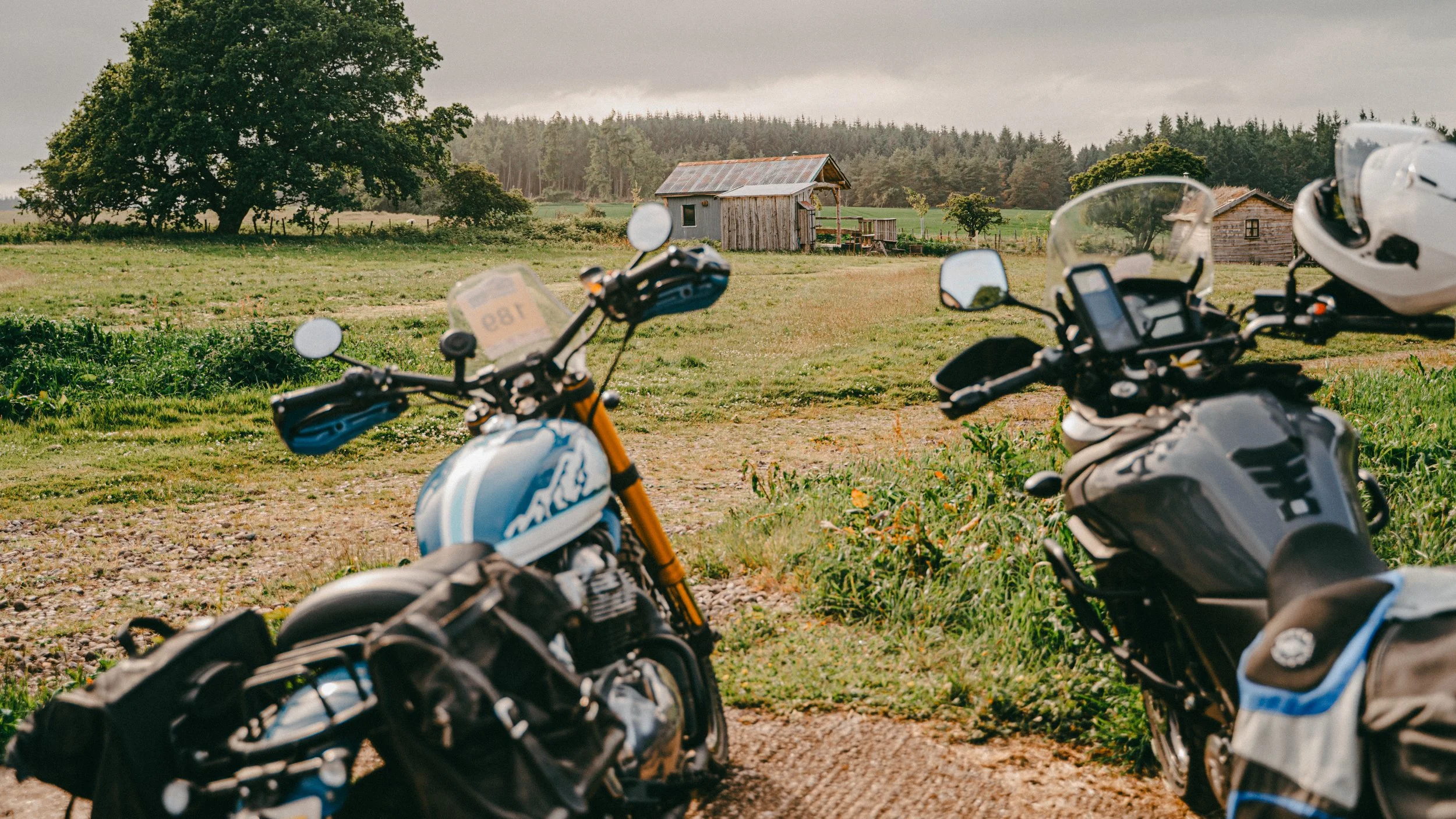 Three motorcycles parked on a grassy area in front of a rural landscape with small wooden houses, trees, and overcast skies.