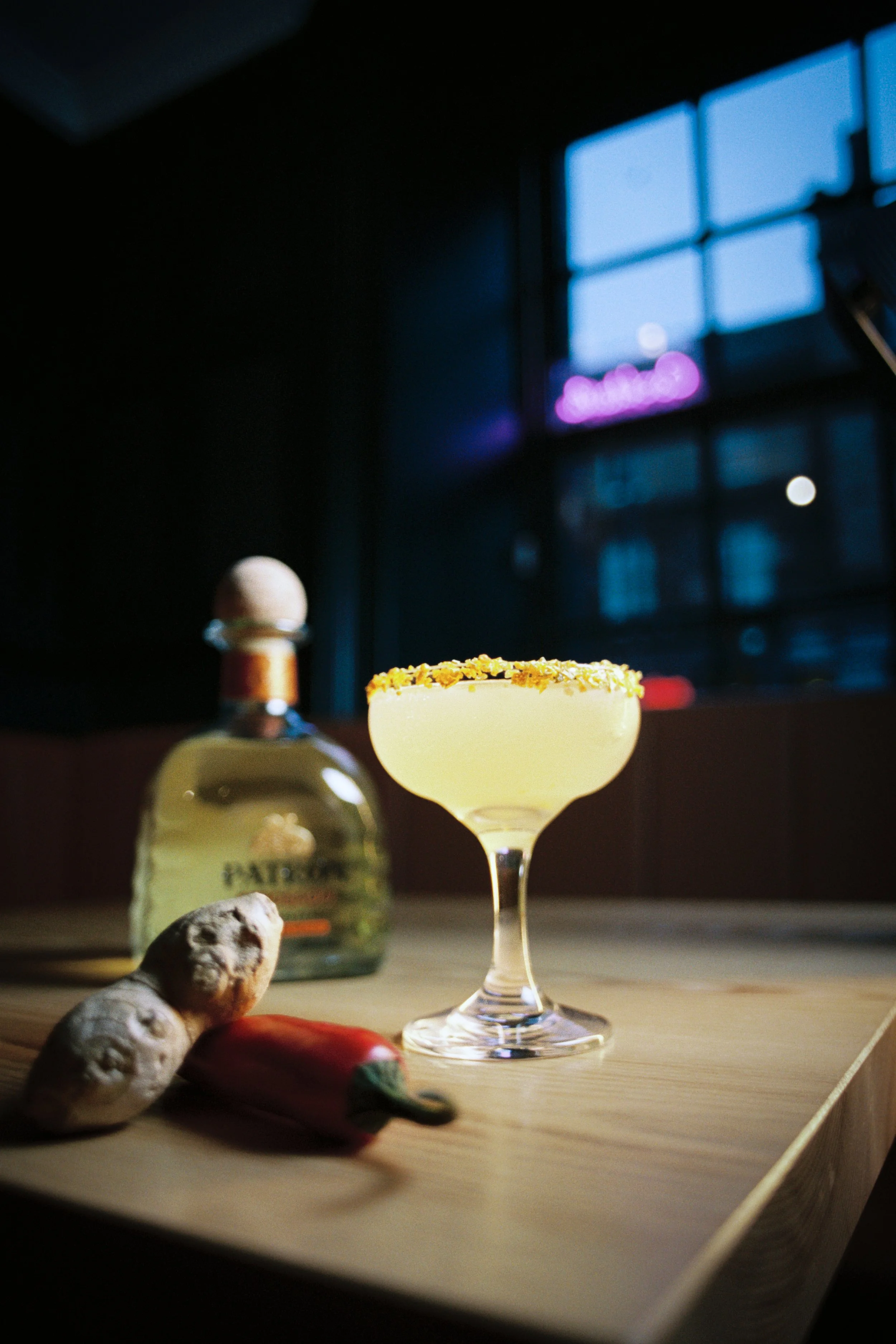 A yellow cocktail with salt on the rim in a coupe glass, on a wooden surface with toppings like ginger, red pepper, and garlic in the foreground. In the background, a bottle of Pyrat rum, a window with blue sky, and a neon sign are visible.