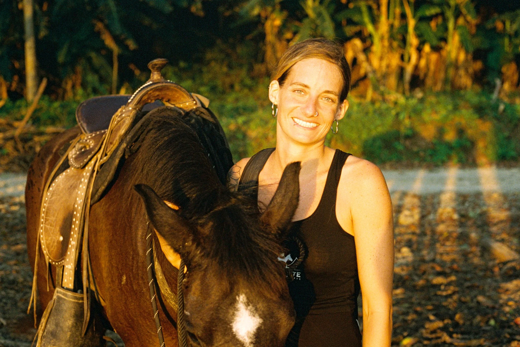 A woman with short brown hair, wearing hoop earrings and a black tank top, is outdoors at sunset, standing next to a dark brown horse with a saddle and tack, in front of a background of trees with autumn leaves.