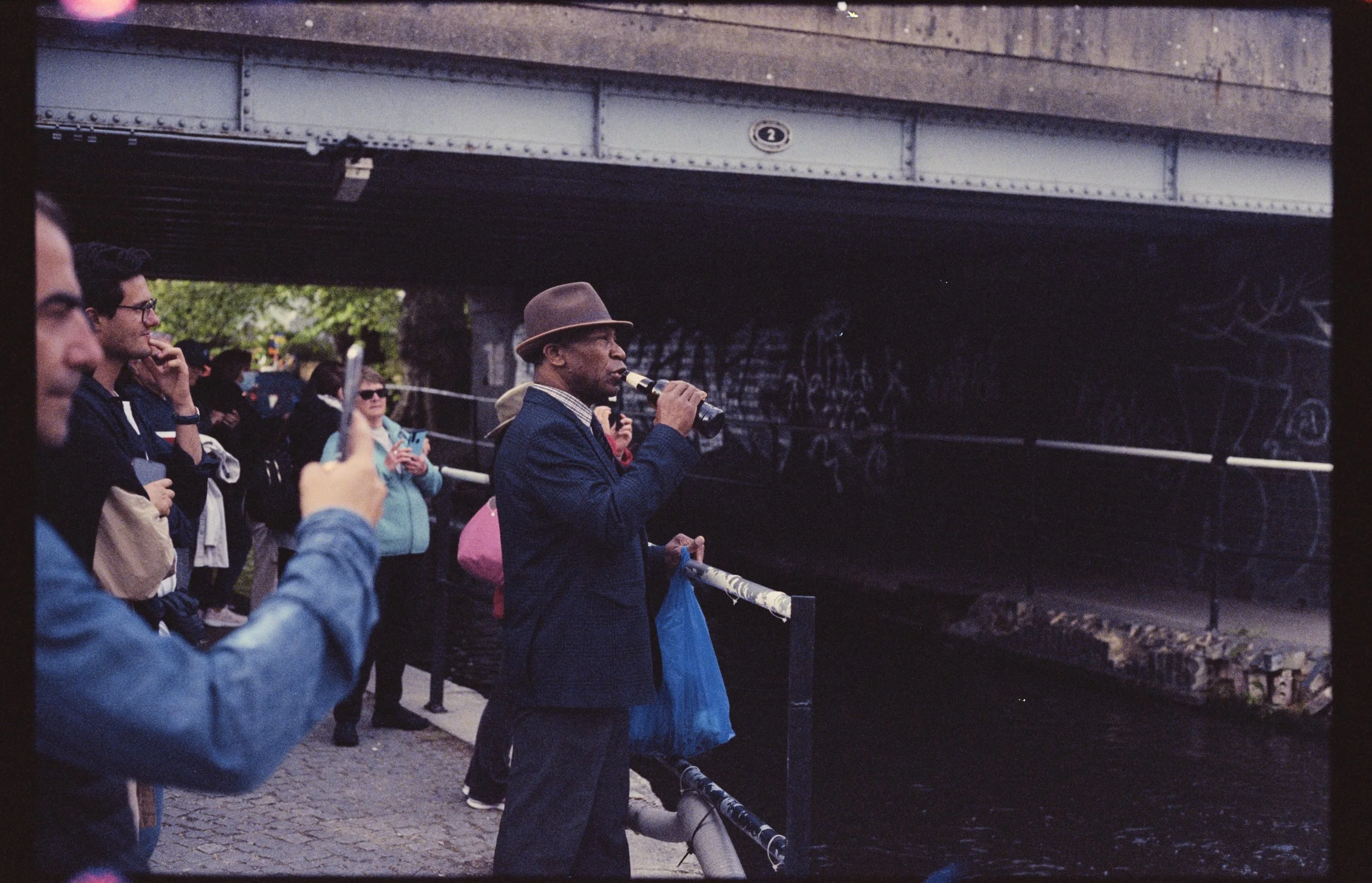 People standing outdoors under a bridge, with some looking at the water and others taking photos, during dusk or evening.