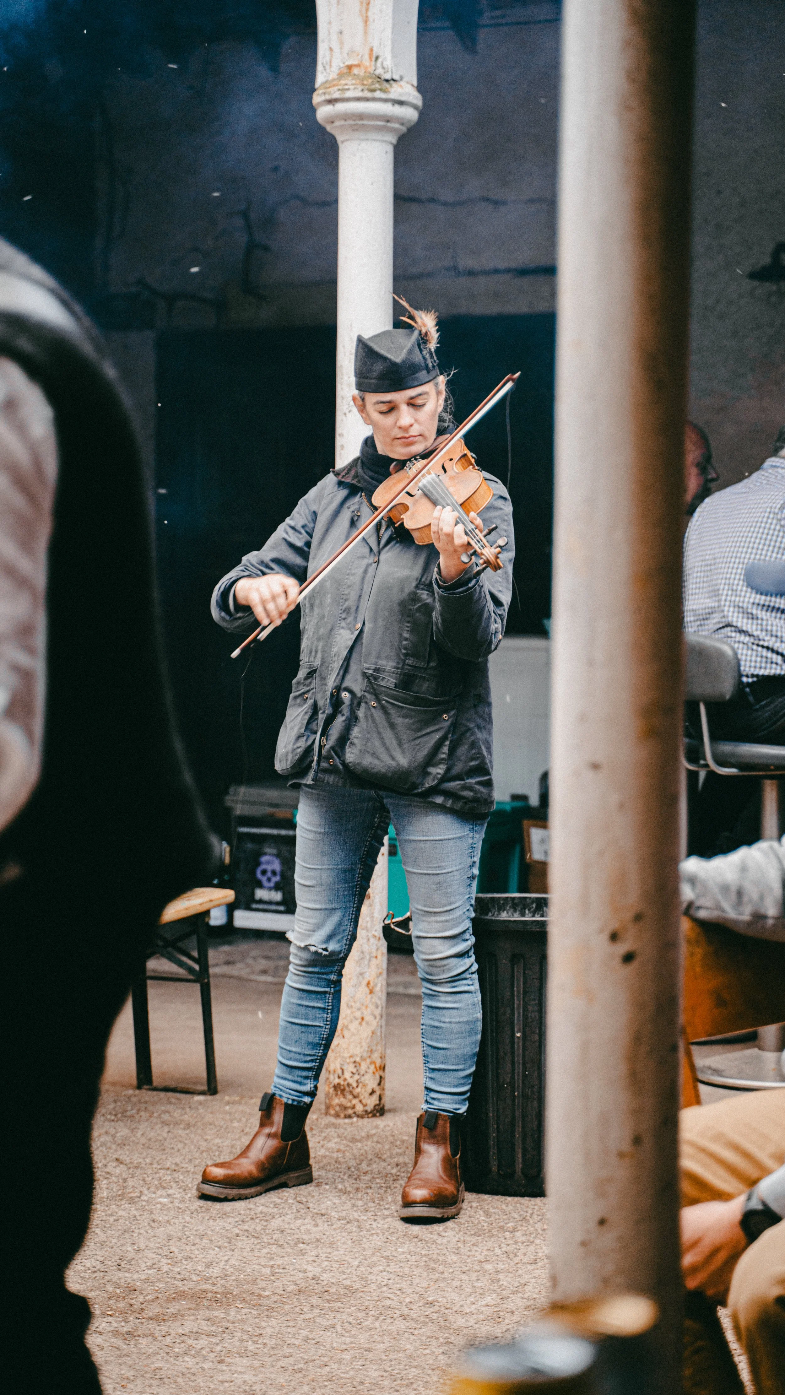A young woman playing the violin indoors, wearing a gray jacket, jeans, and brown boots, with a black hat decorated with a feather.