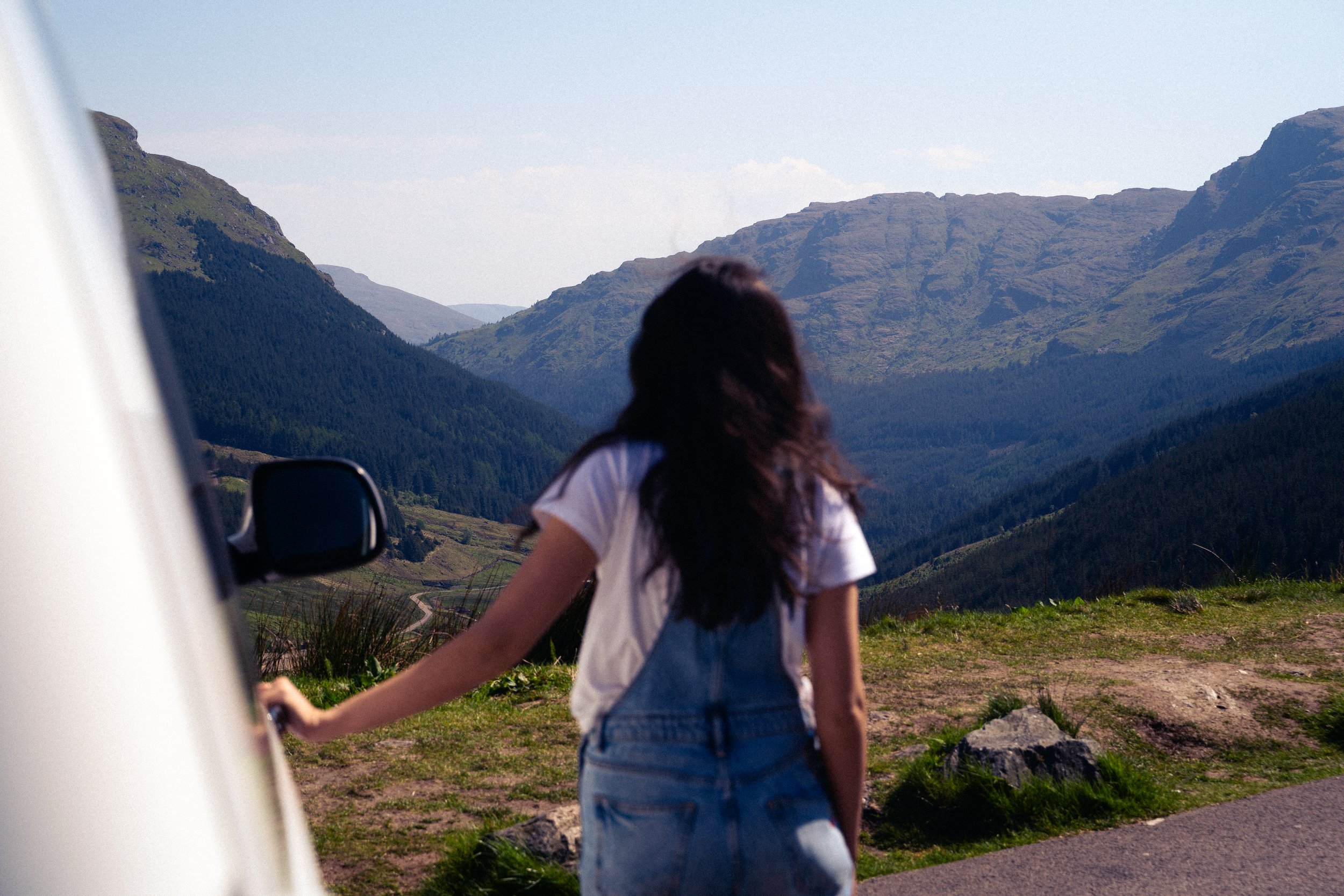A woman with long, dark hair, wearing a white t-shirt and denim shorts, holding a bicycle handlebar, standing in a scenic mountainous landscape with green hills and rugged terrain under a clear sky.