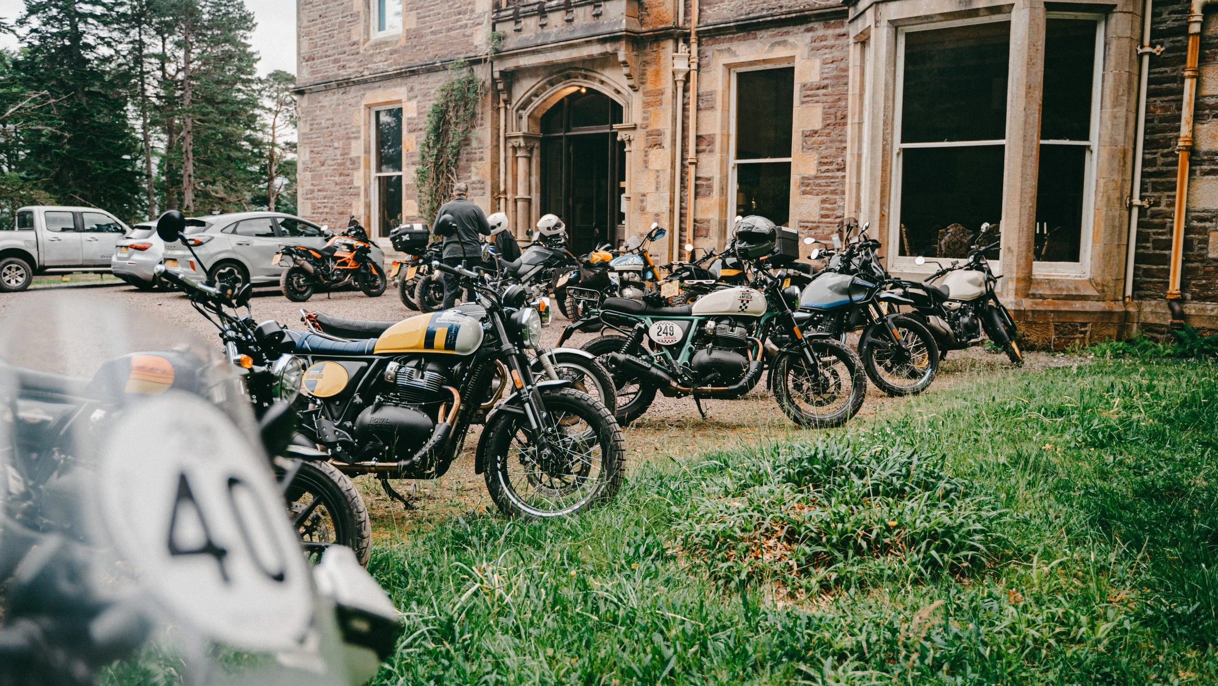 Multiple motorcycles and cars parked outside a historic brick building with large windows, with a person standing near the motorcycles.