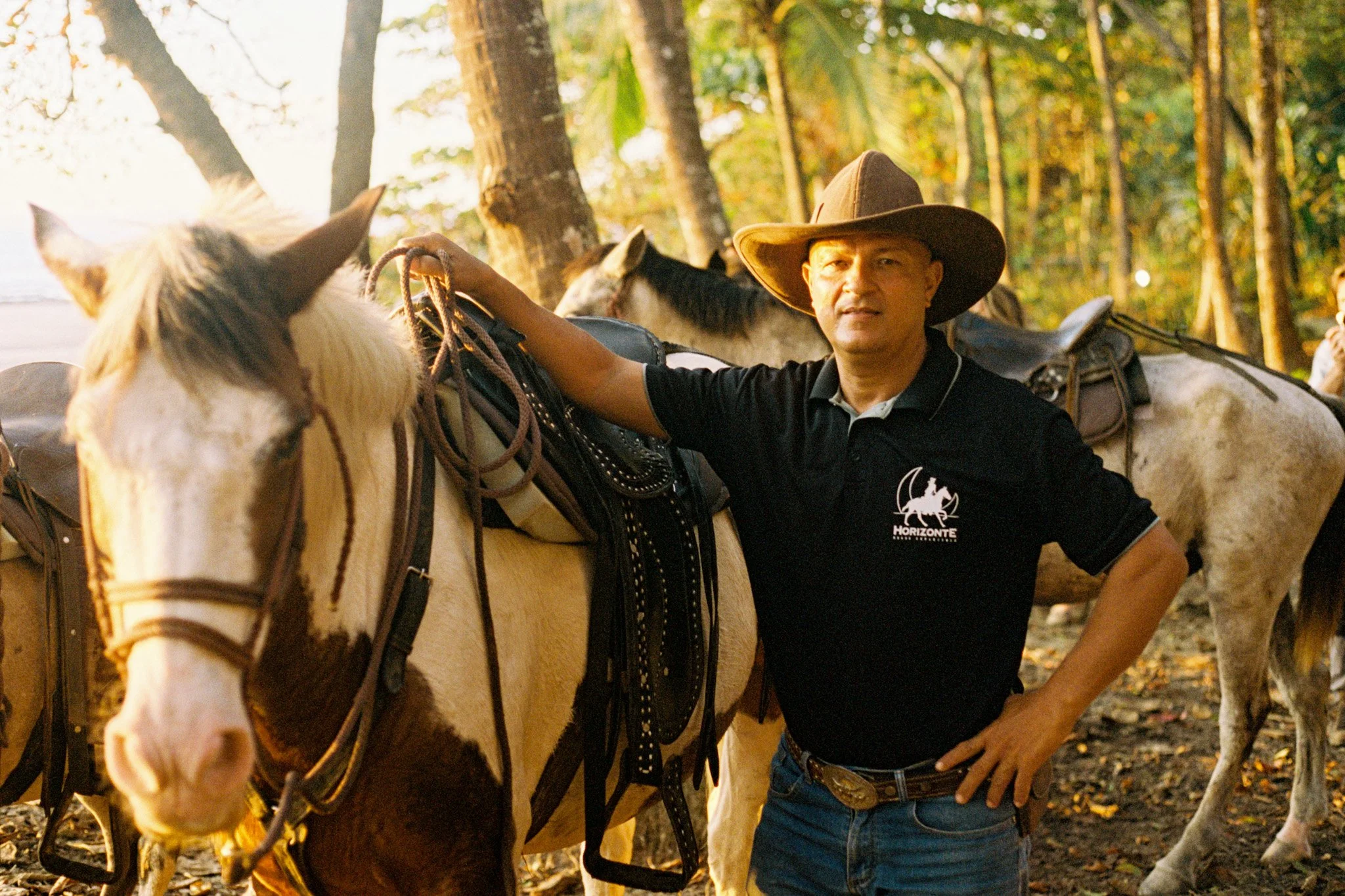 A man wearing a cowboy hat and black shirt with a horizonte logo stands next to a white horse with brown markings in a wooded area during autumn. The man is holding the horse's reins and looking at the camera.