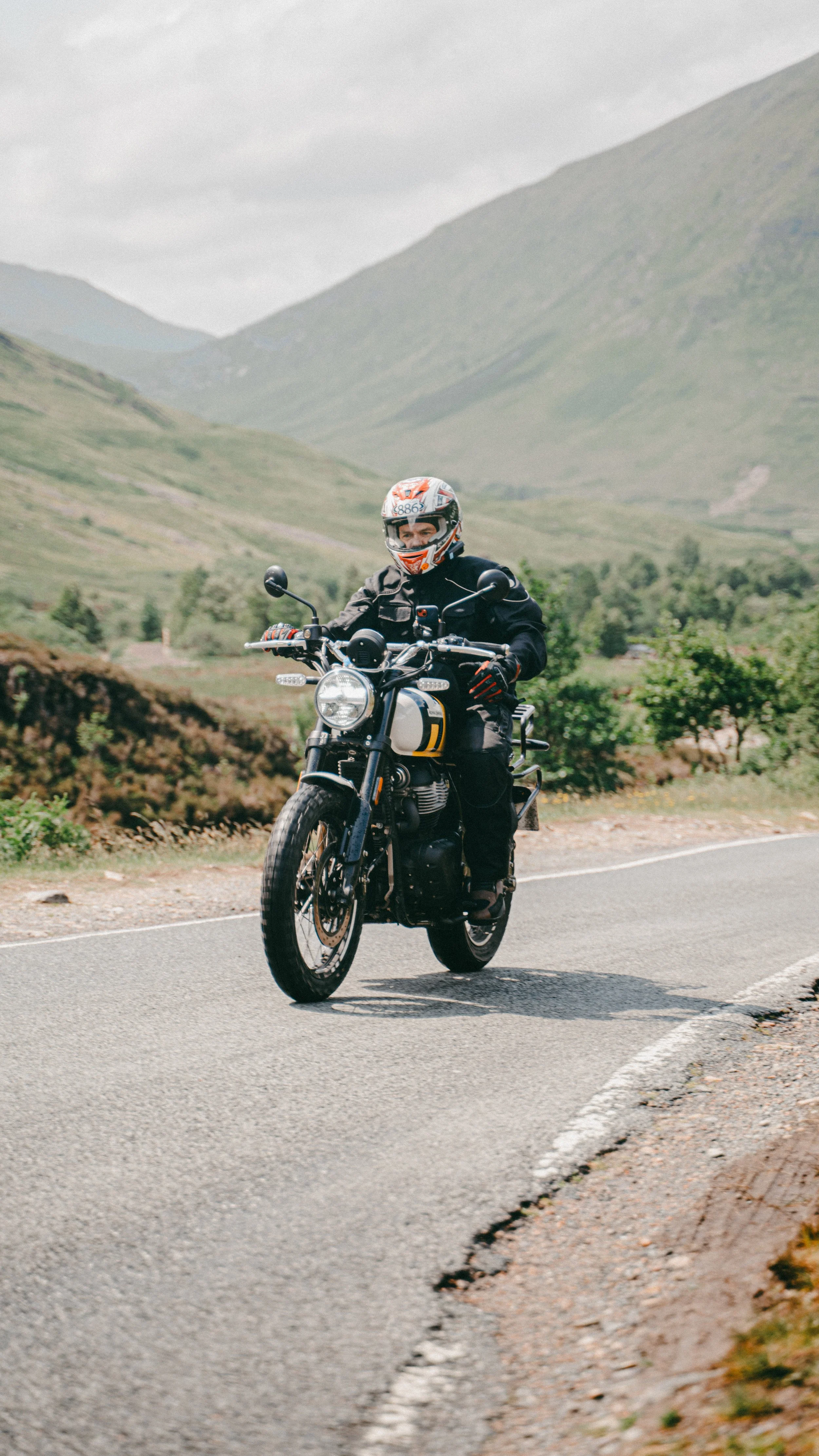 A person riding a motorcycle on a mountain road with green hills and a cloudy sky in the background.