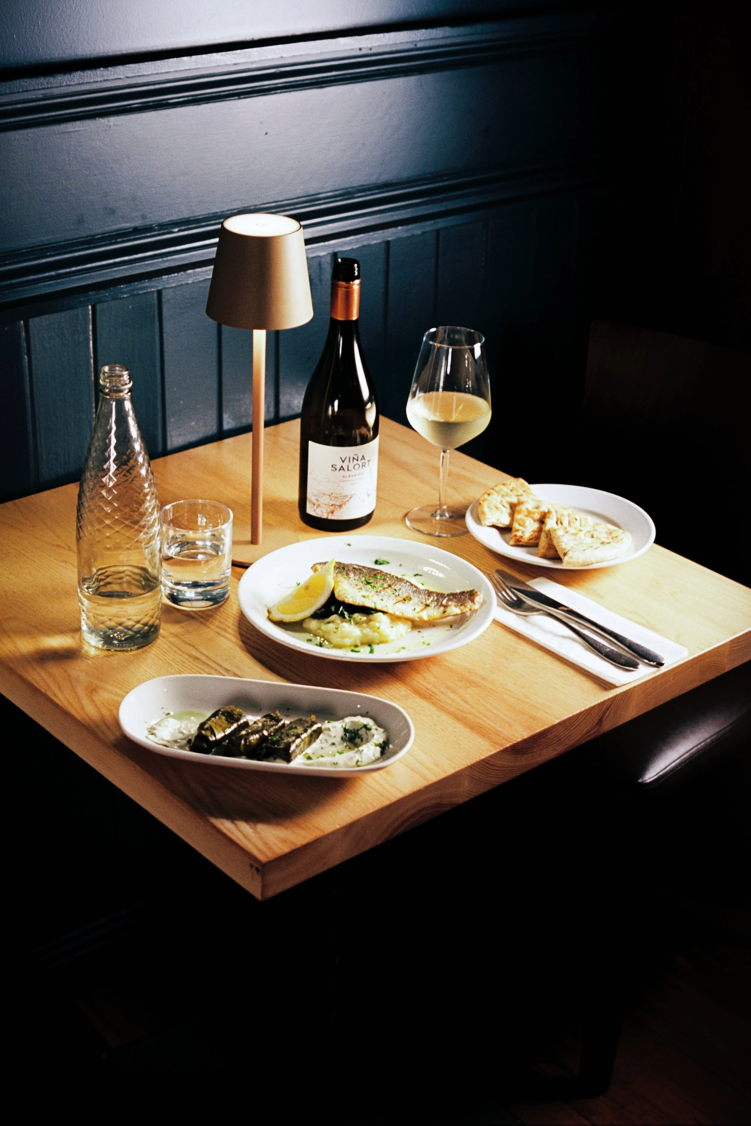 A wooden dining table with a glass of white wine, a bottle of wine labeled 'Vina Salori,' a white plate with a fish dish garnished with lemon, a bowl of grilled vegetables, a small lamp, a bottle of water, and a plate of bread on the table. The backg