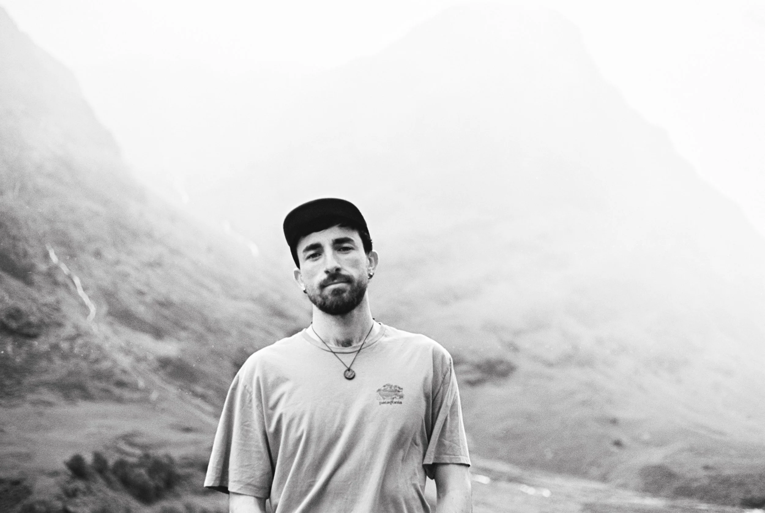 A young man with dark hair and a beard standing outdoors with mountains and a cloudy sky in the background, wearing a baseball cap, t-shirt, and necklace.