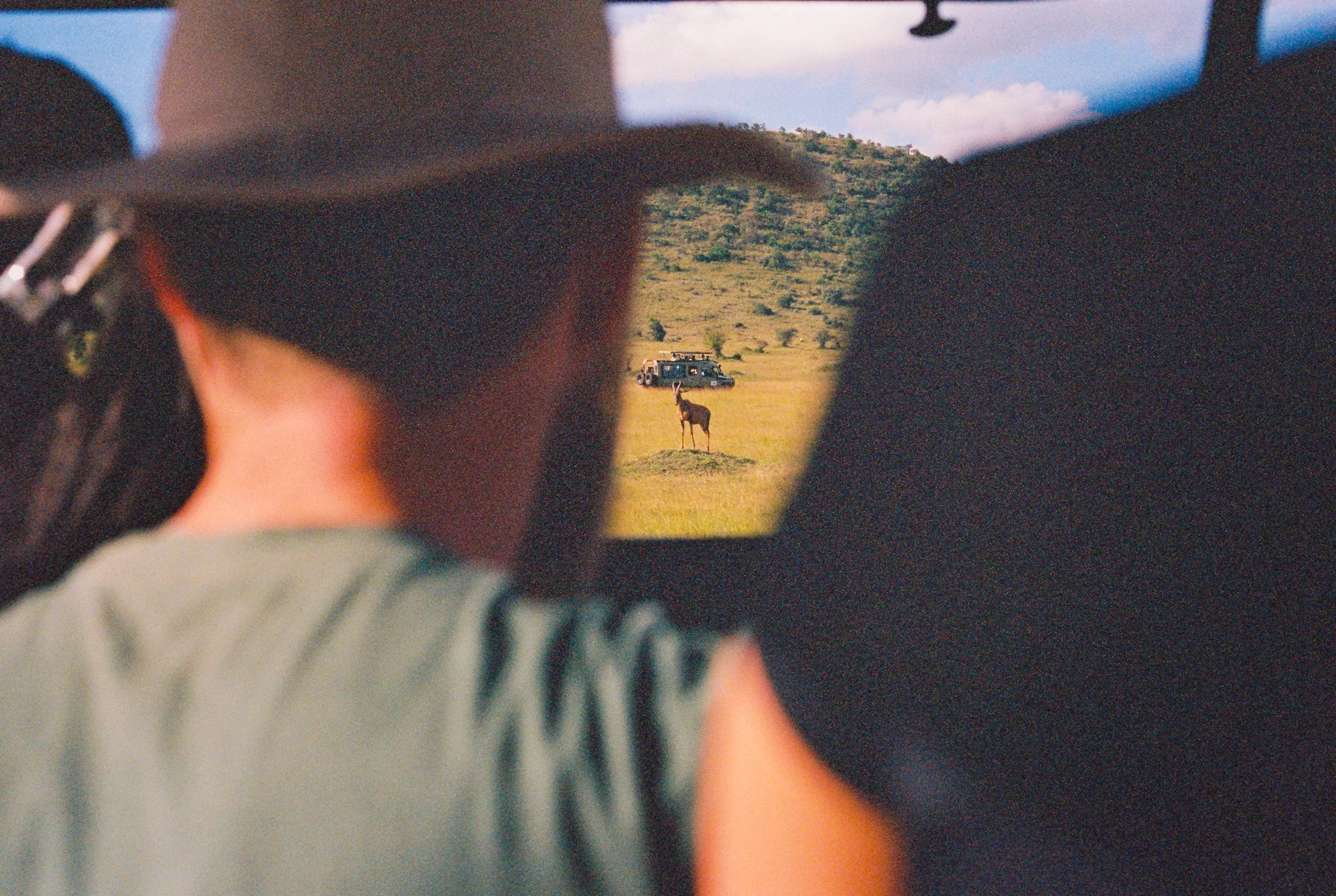 View through a mirror showing a safari scene with a vehicle, a giraffe, and greenery, with the person taking the photo partially visible.
