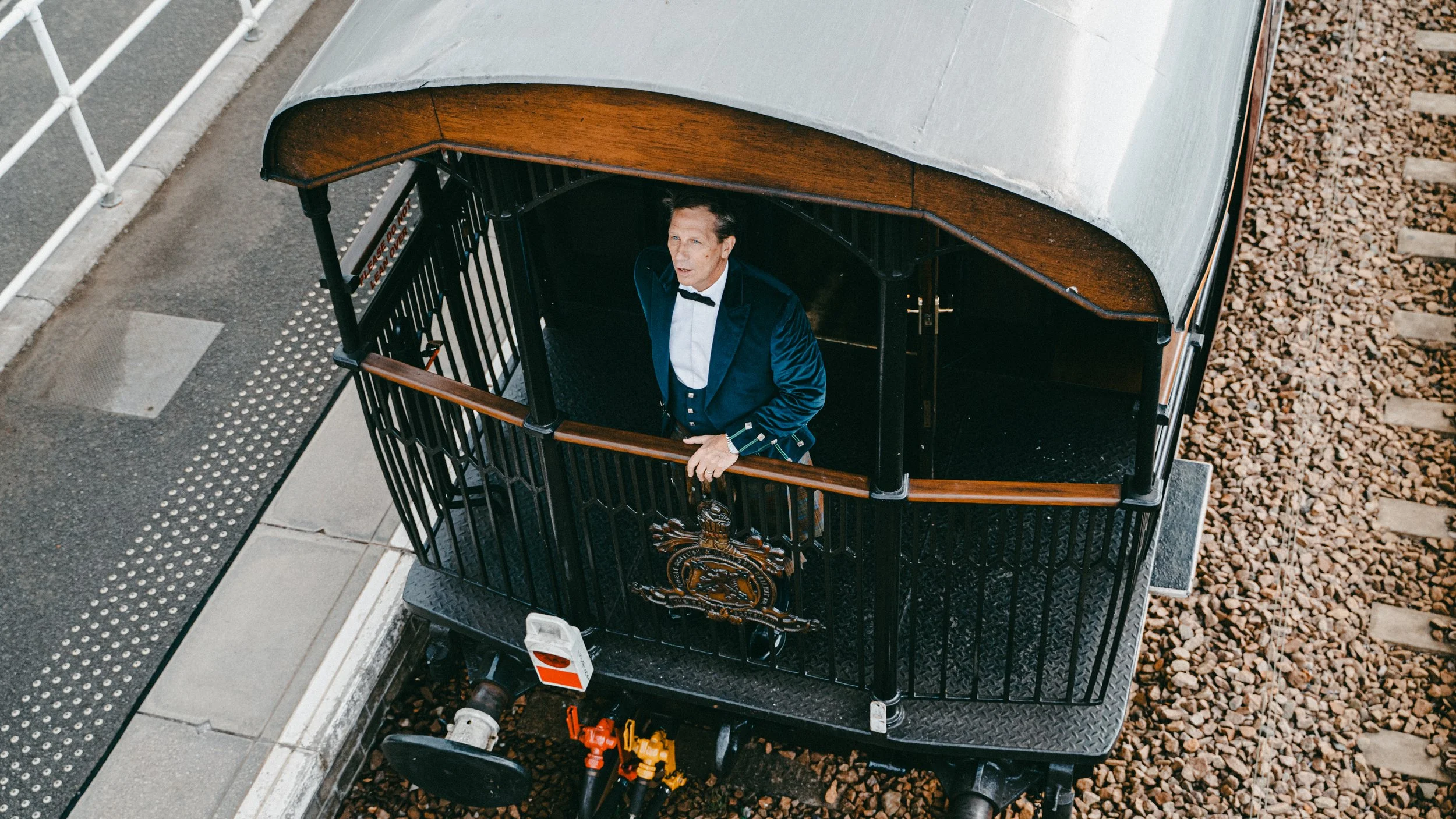 A man in formal attire, including a tuxedo with a bow tie, standing inside a black, vintage-style trolley or carriage on railroad tracks, looking up. The carriage has a wooden roof and a decorative emblem on the front.