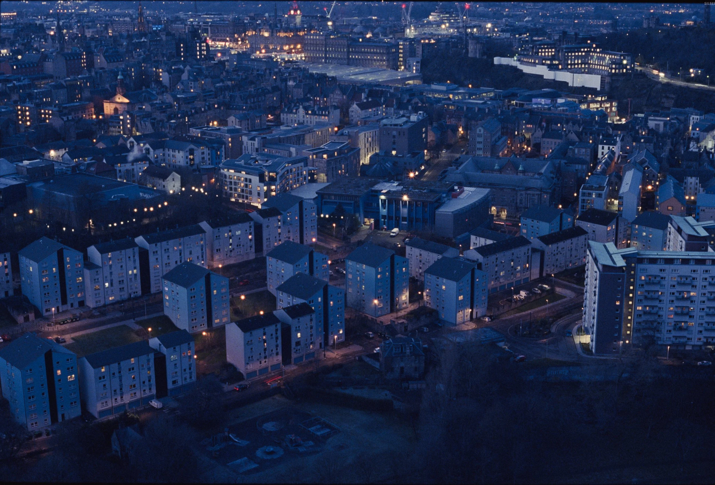 A cityscape at dusk with buildings, streets, and lights illuminated against a darkening sky.