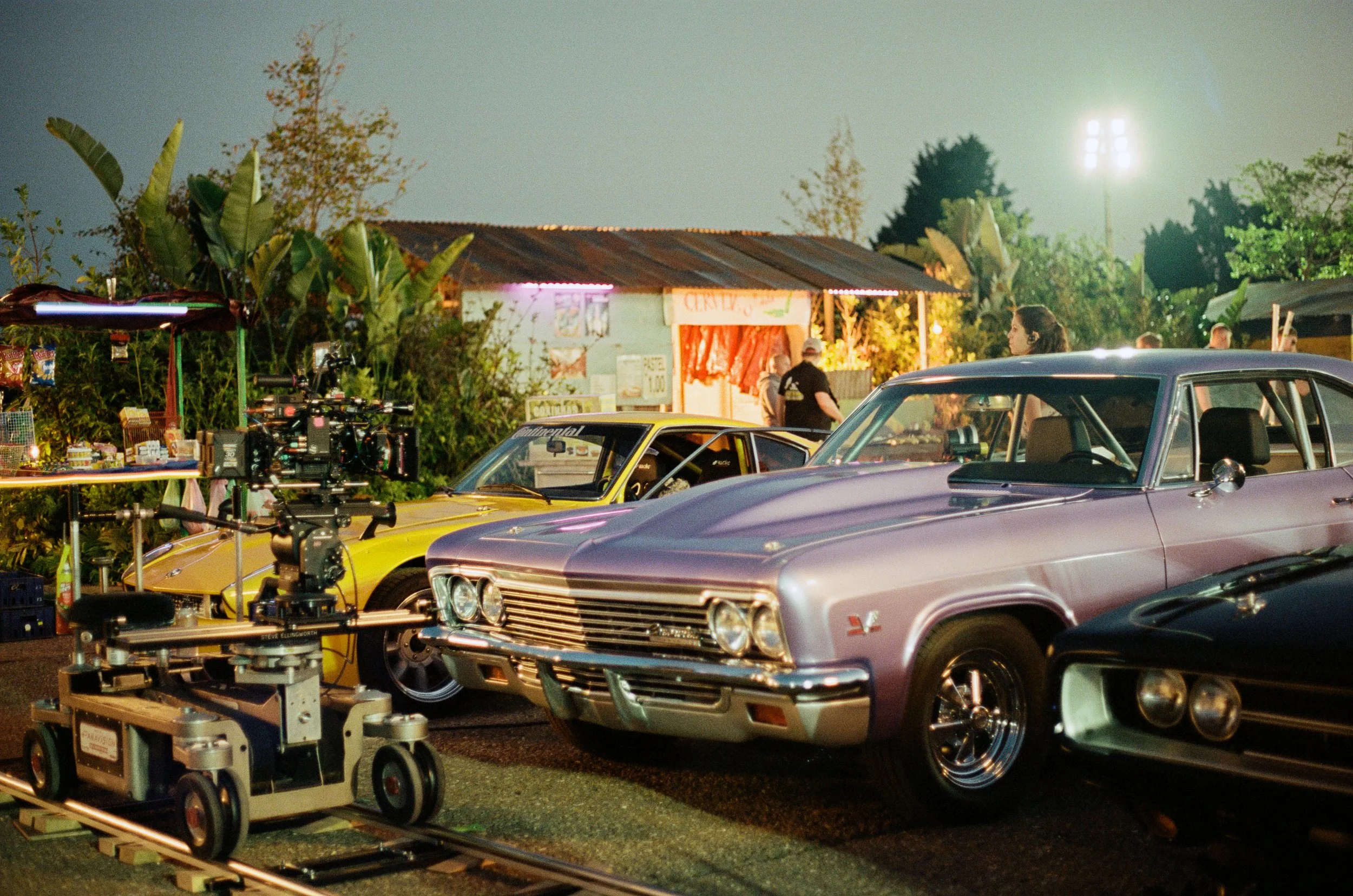 Nighttime scene featuring vintage cars parked outdoors, a film set with camera equipment, and a small building with a sign, surrounded by trees, under bright floodlights.