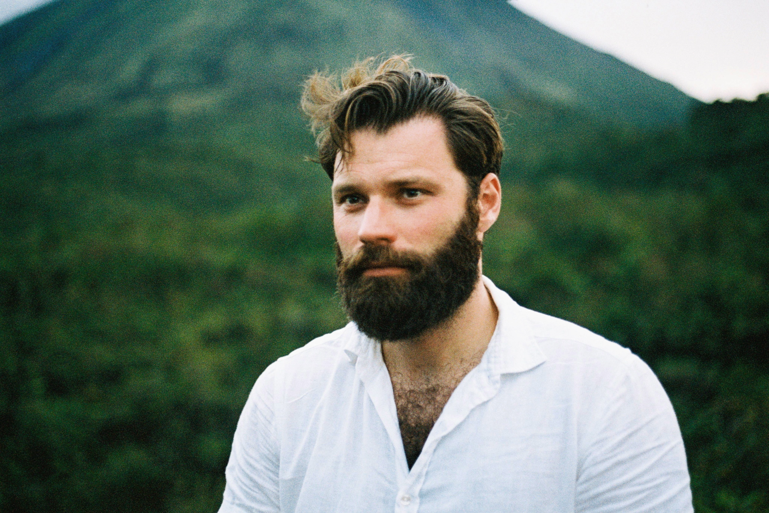 A man with dark hair and a full beard wearing a white button-up shirt, standing outdoors in front of a mountain with green trees and cloudy sky in the background.