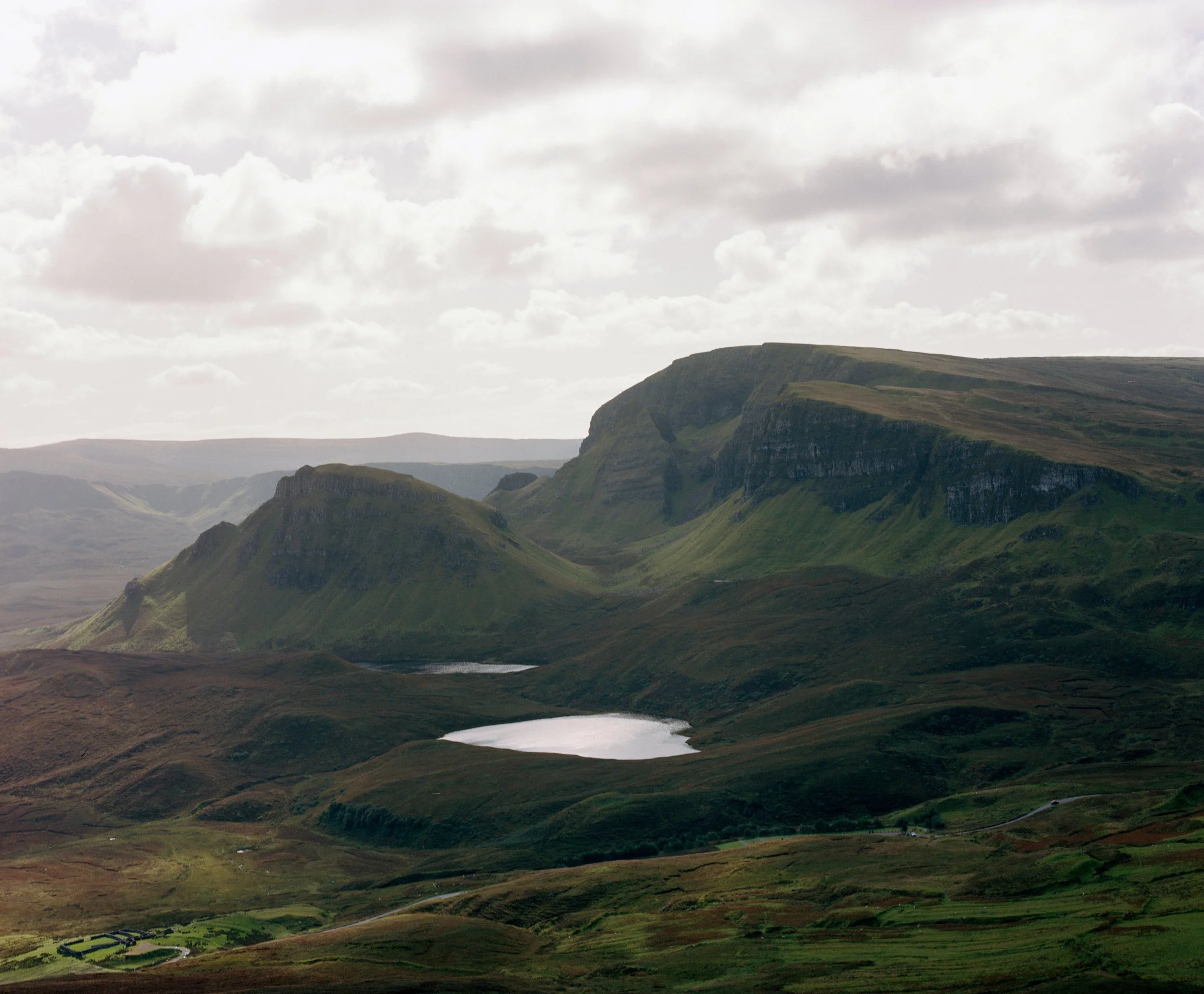 A scenic landscape of rugged green hills and cliffs with patches of water, cloudy sky overhead.