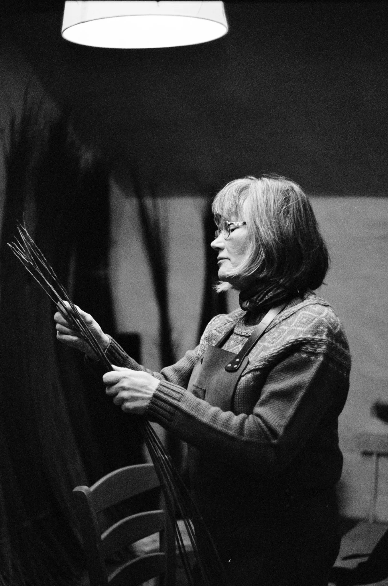 A woman with shoulder-length hair, glasses, and a sweater, standing in a room while holding a bunch of thin sticks or brushes, possibly in an art or craft setting.