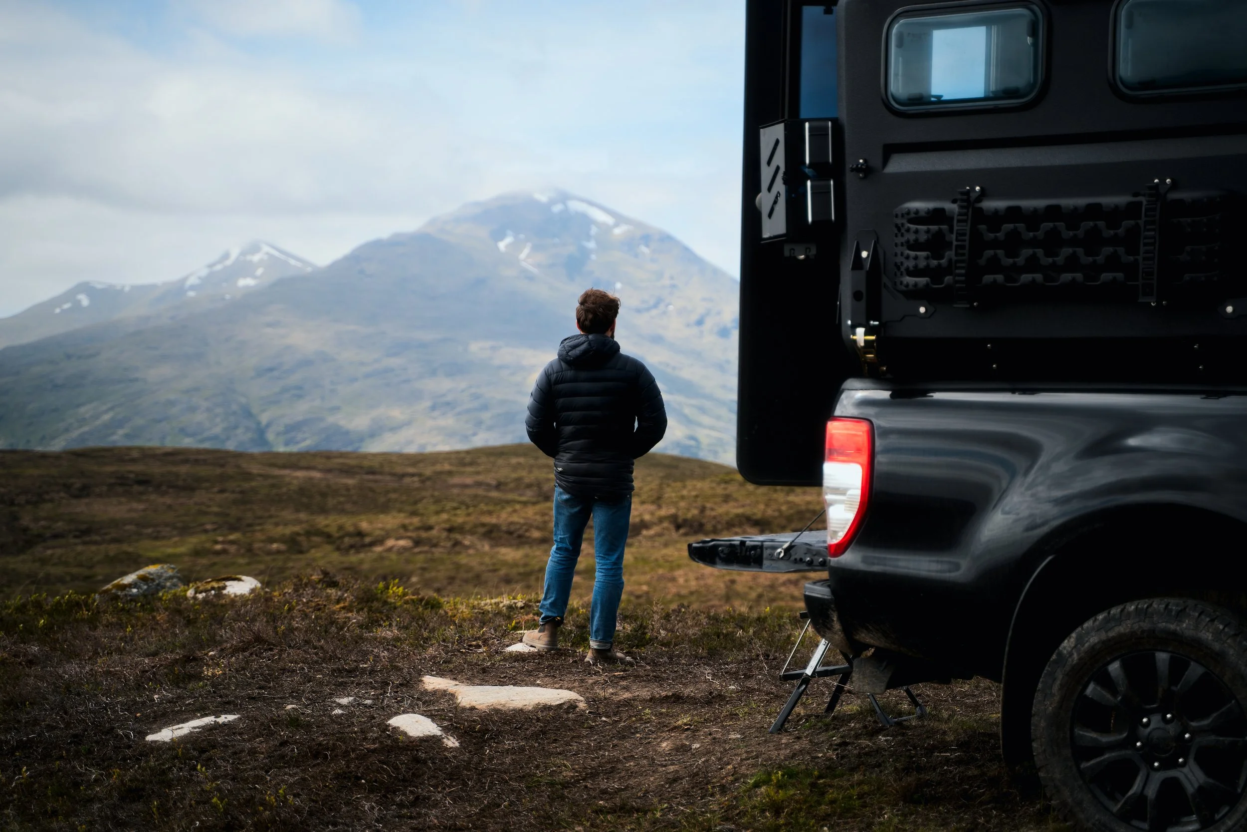 A person standing outdoors in front of a mountain landscape with snow patches, next to a black pickup truck with camping gear. The person is wearing a black jacket and jeans, facing away from the camera, with hands in pockets.