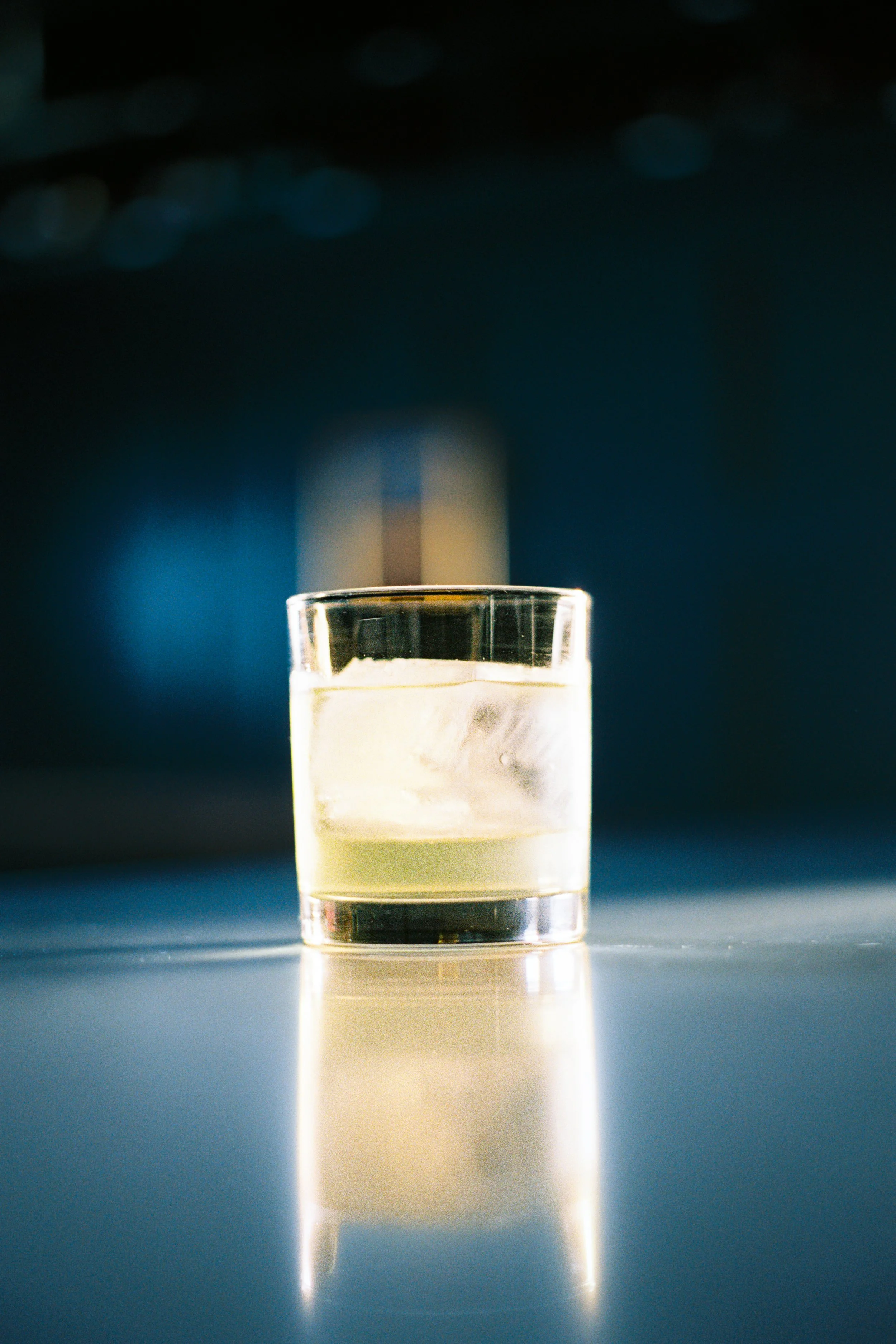 A close-up of a glass with a yellowish transparent liquid, probably whiskey, against a dark background with blue highlights and bokeh effects.