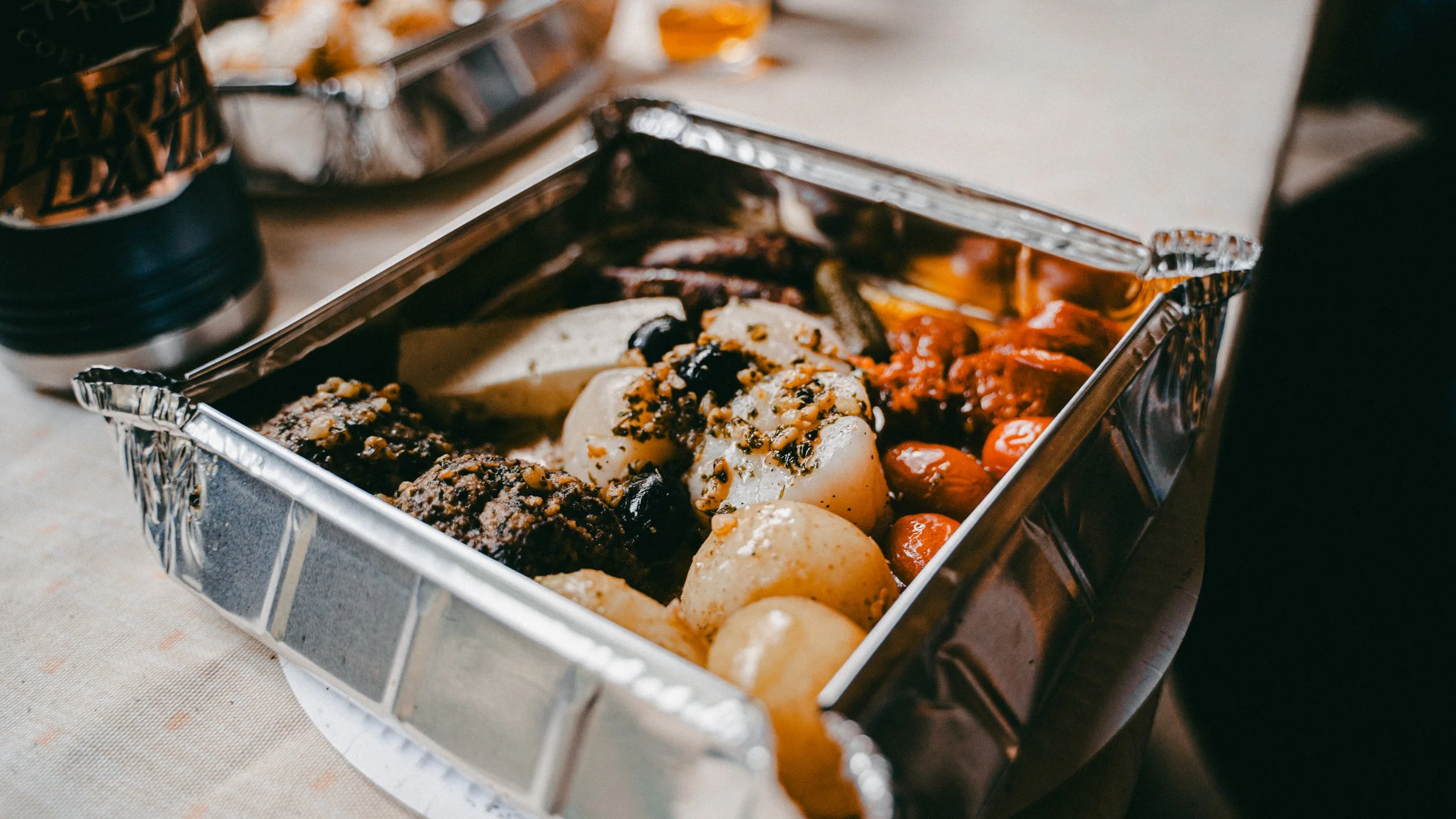 A metal buffet tray with baked potatoes topped with cheese and herbs, grilled chicken pieces, cherry tomatoes, and various cooked vegetables.