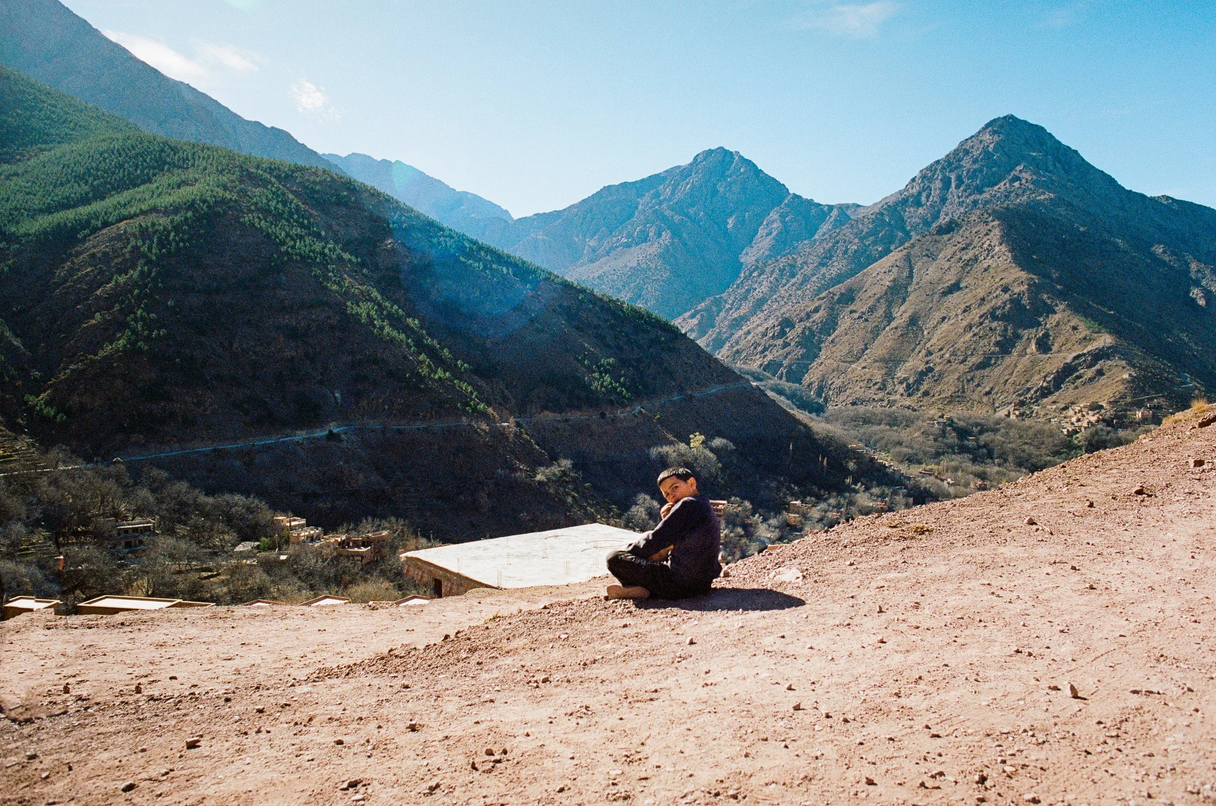 A child sits on a ledge overlooking a mountainous landscape with small village buildings at the valley floor and green forested mountains under a clear blue sky.