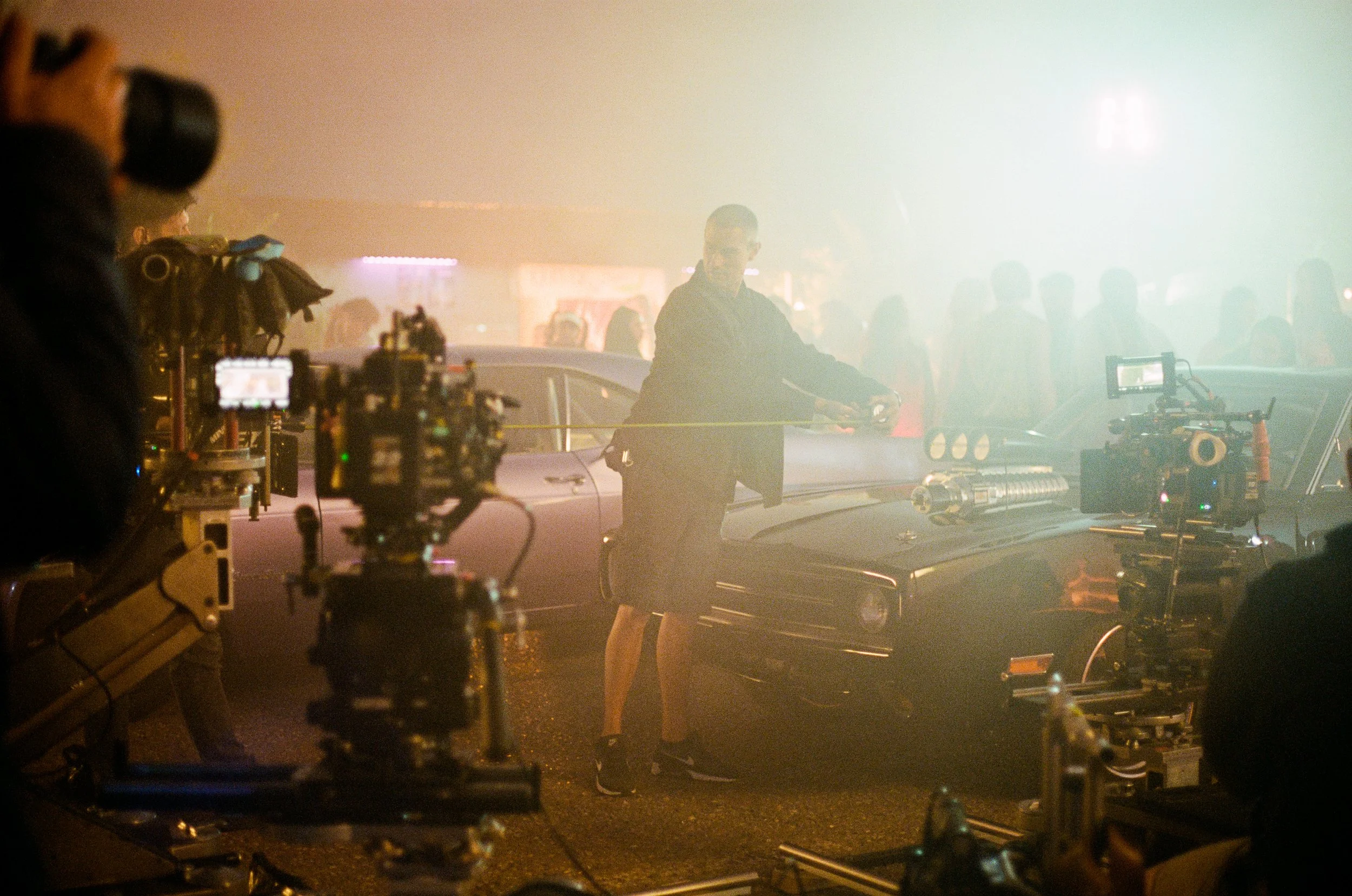 Behind-the-scenes shot of a film production, showing camera equipment and a man with glasses and a black jacket in a foggy or smoky environment, possibly on a city street at night.