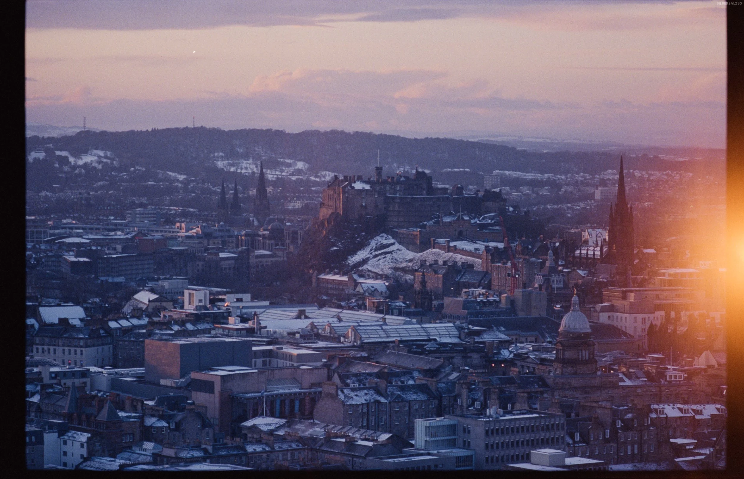 A cityscape view of a historic fortress on a hill, surrounded by buildings, with snow on rooftops and distant hills, during sunset with pink and purple sky.