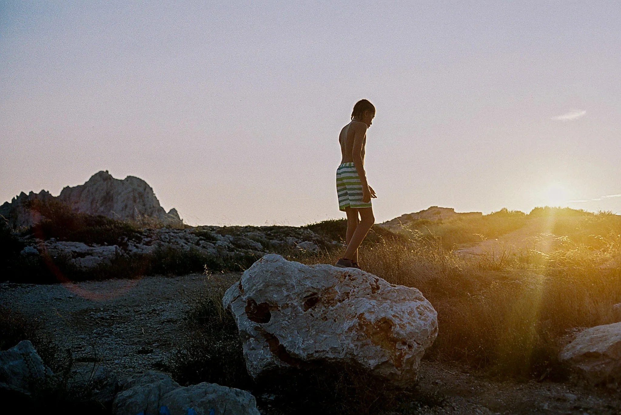 Person walking on a rocky terrain during sunset or sunrise with mountains in the background.