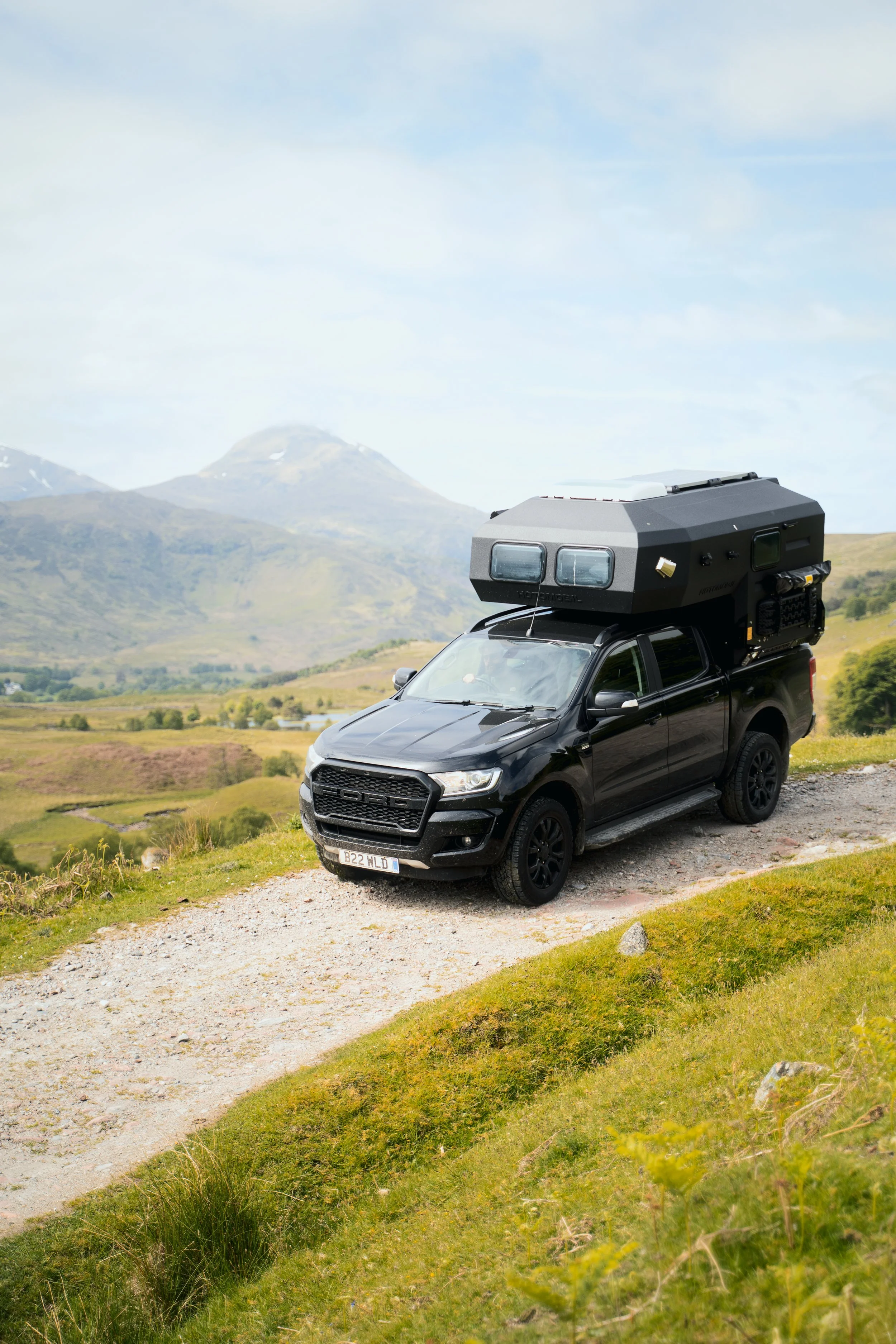 A black pickup truck with an attached rooftop camper parked on a grassy and gravelland landscape, with hills and mountains in the background under a partly cloudy sky.