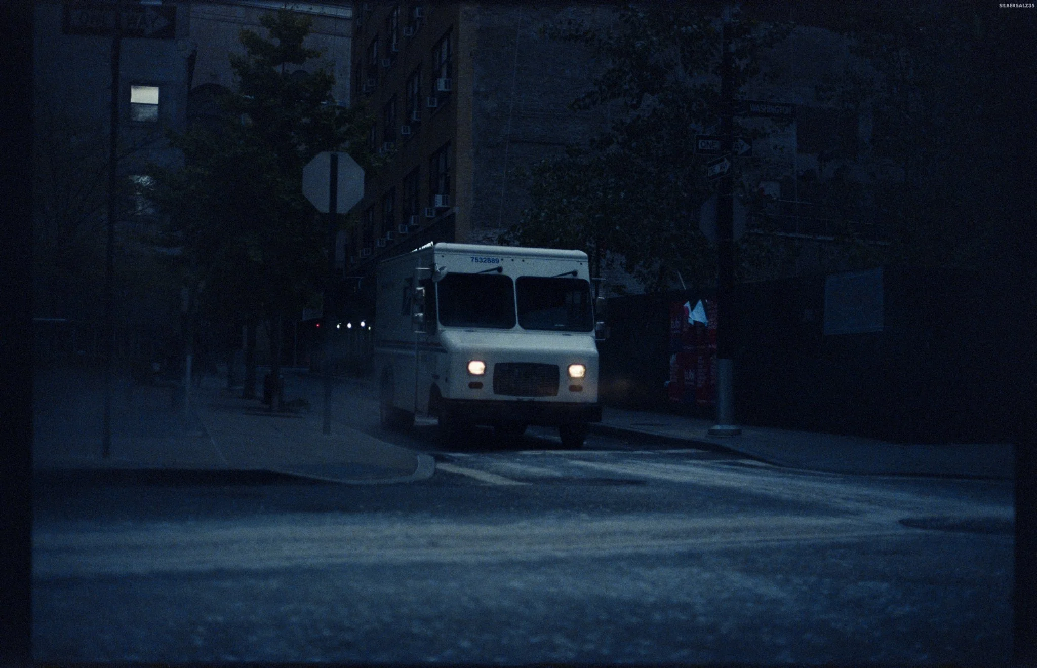 A white delivery truck on a city street at night, with buildings and trees in the background, and a dark sky overhead.