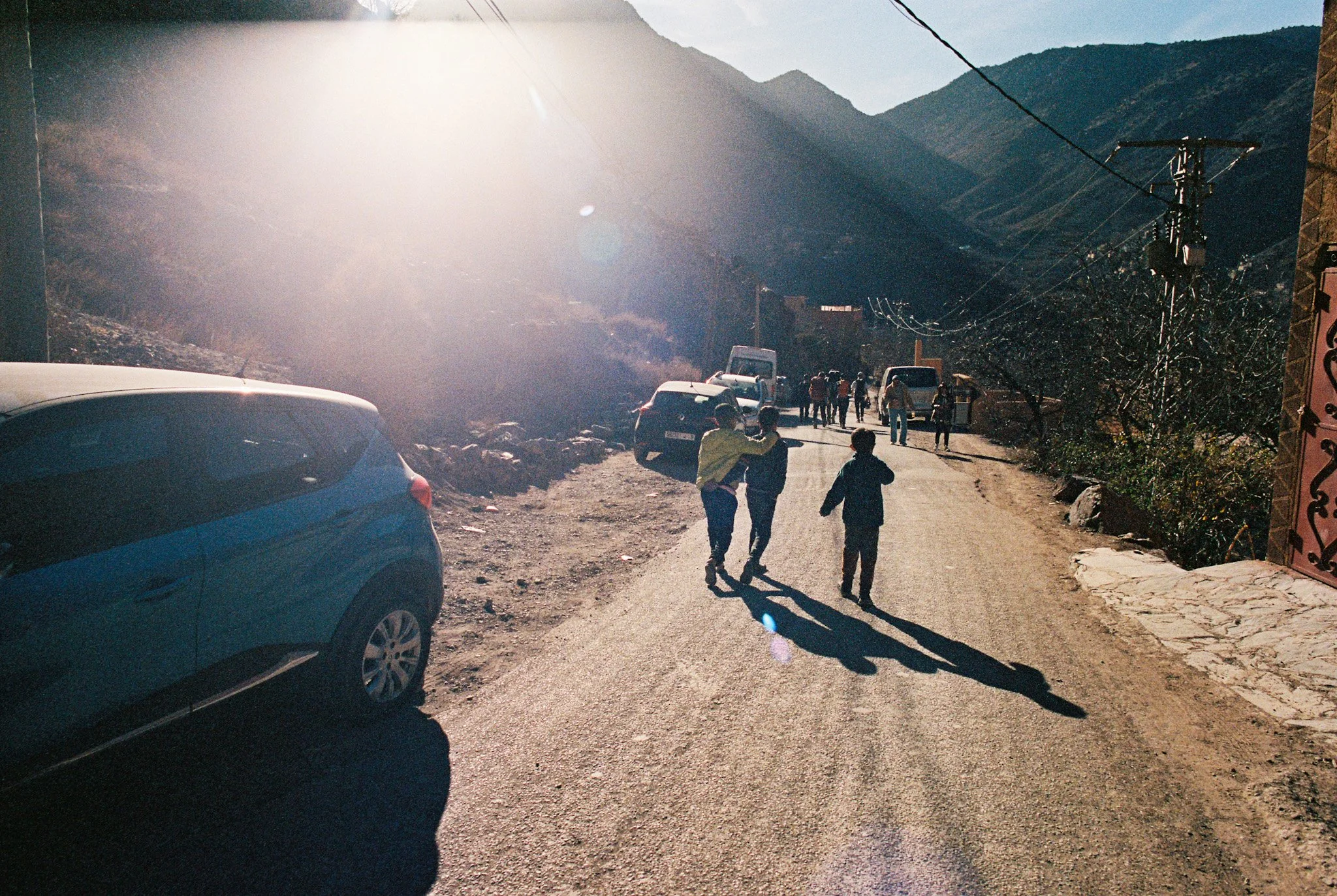 A mountain road scene with cars parked along the side, a group of people walking, and mountains in the background under a bright sun.