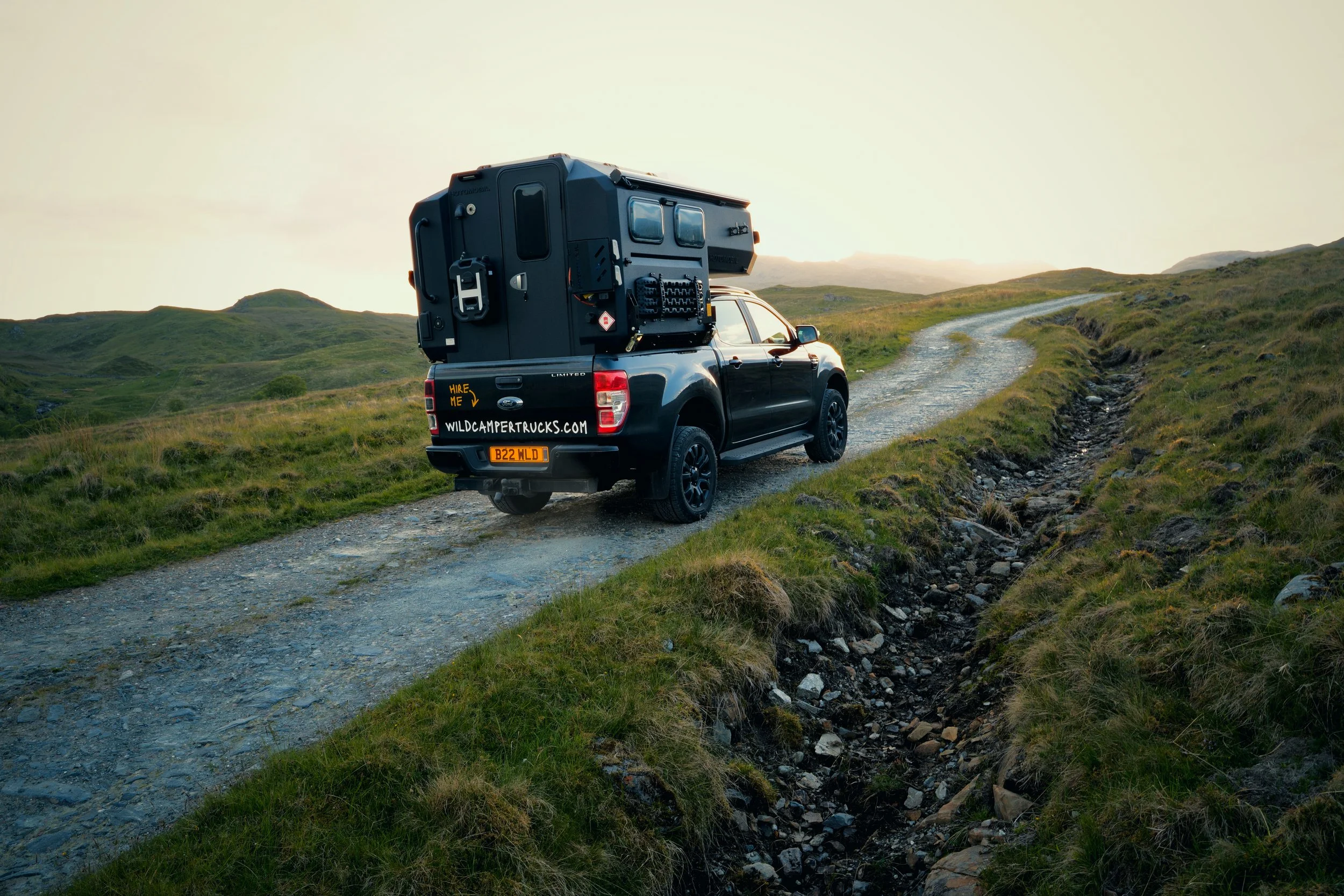A black pickup truck with a large camper attached, driving on a winding dirt road through green hills at sunset.