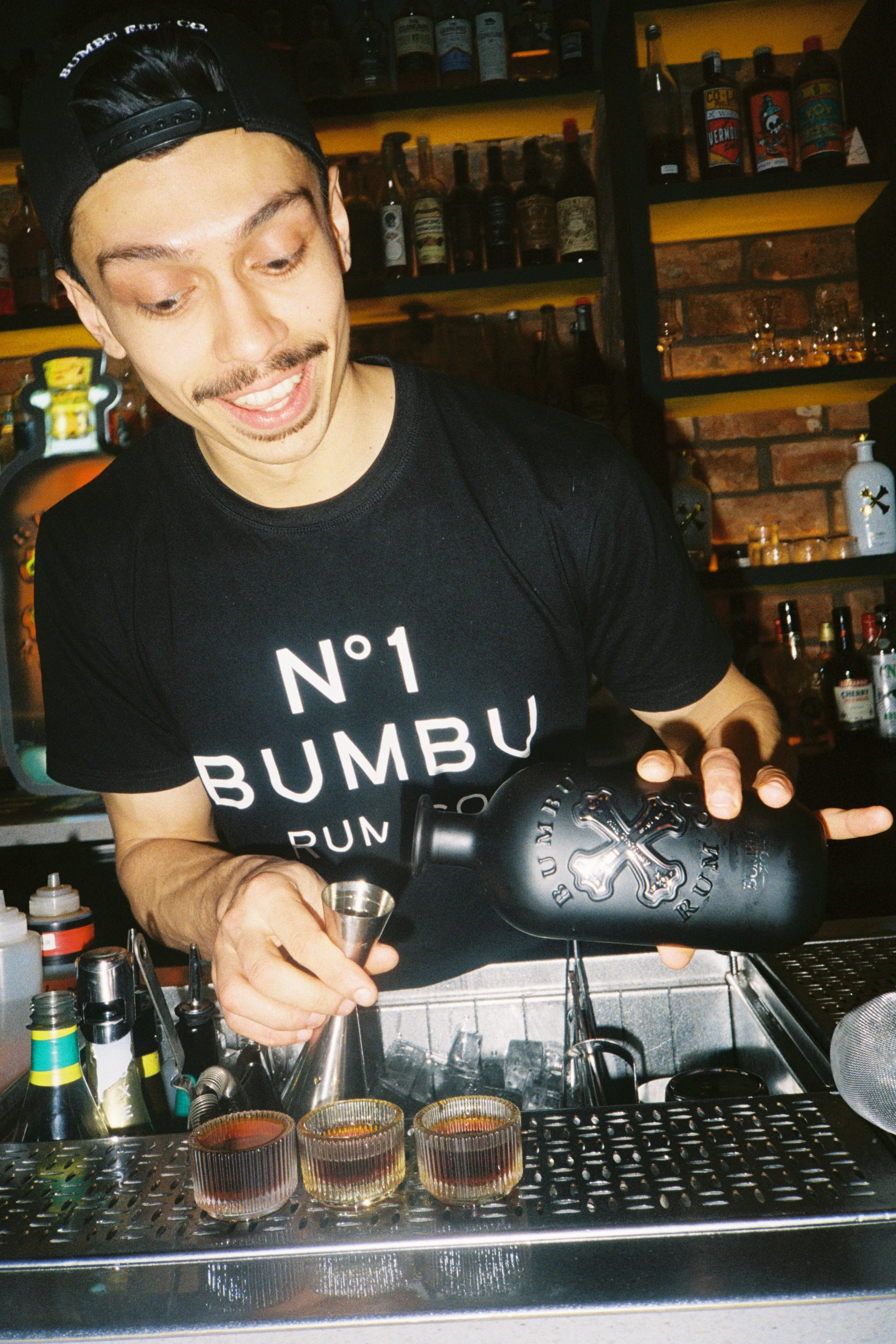 A bartender wearing a black cap and black t-shirt preparing cocktails behind a bar. Several glasses with drinks are in front, and bottles of alcohol and mixers are on shelves in the background.